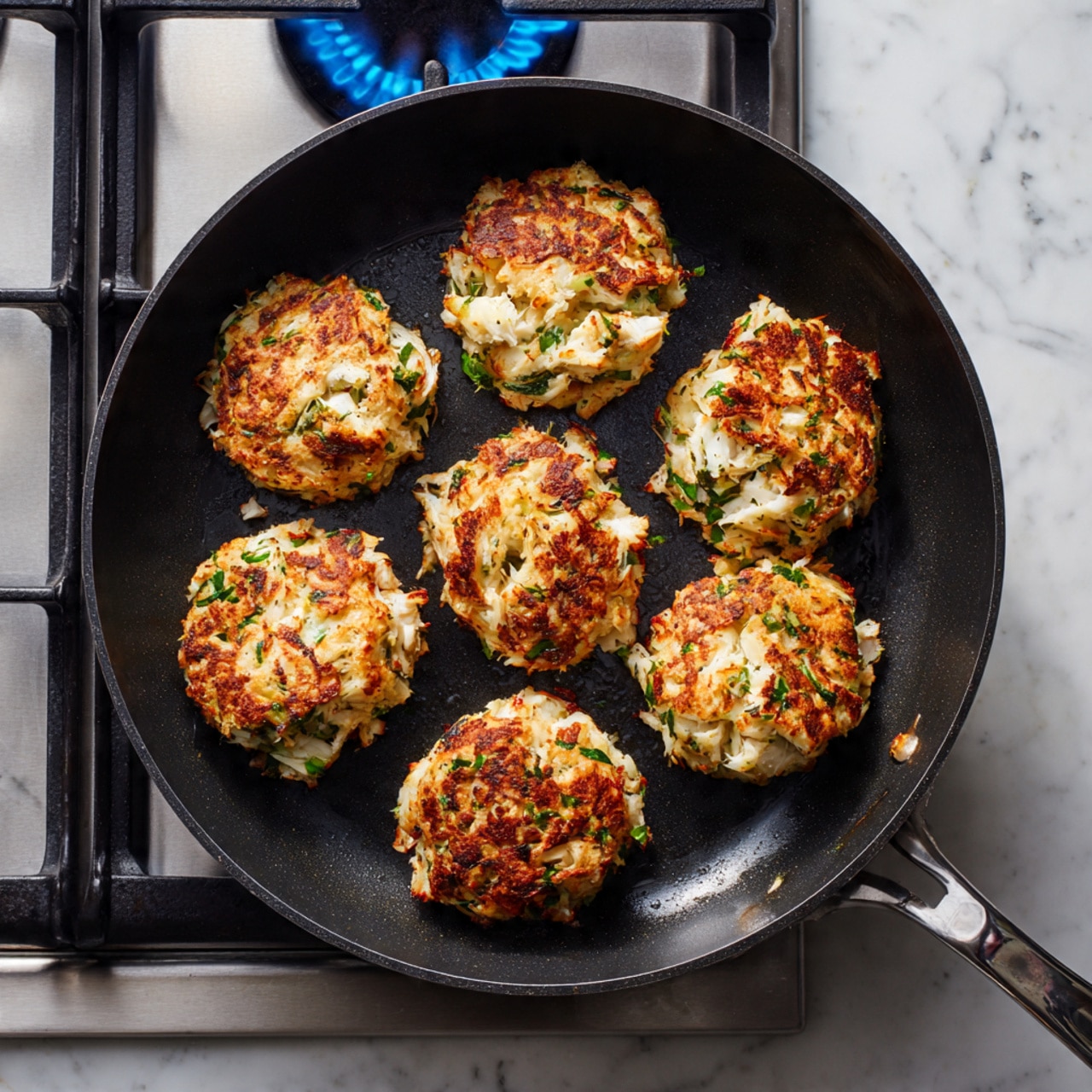 A black frying pan sits on a gas stove with blue flames beneath. Inside the pan are six golden brown crab cakes, each roughly round and about two layers thick, with a crispy textured top layer showing bits of green herbs and white crab meat. The cakes have a slightly uneven shape with visible chunks of crab and parsley mixed throughout, creating a mix of creamy white, green, and golden brown colors. The stove is set on a white marbled surface that adds a clean and bright feel to the scene. photo taken with an iphone --ar 4:5 --v 7