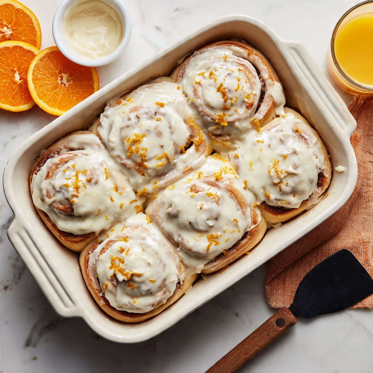 A soft, round cinnamon roll with three visible layers: the bottom fluffy dough layer in light golden yellow, a middle swirled dough with a hint of cinnamon color, and a smooth, creamy white glaze layer on top with small bits of orange zest. The roll is on a white plate on a white marbled surface. A golden fork held by a woman's hand with light purple nail polish lifts a piece from the front. In the background, there is a white baking dish and some orange slices on the marble surface, with a small blue vase of bright yellow flowers nearby. A slightly folded white cloth with blue stripes lies on the bottom right. Photo taken with an iphone --ar 4:5 --v 7