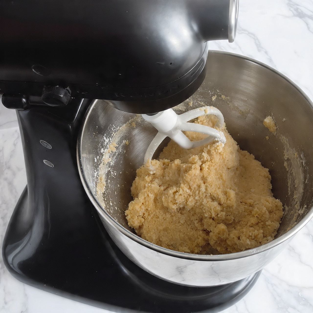 A close-up of a metal mixing bowl attached to a black stand mixer filled with light brown dough being mixed by a white paddle attachment. The dough has a thick, slightly grainy texture with some parts sticking to the sides of the bowl. The background is a white marbled texture, and the mixer has the brand name “Kitchen” visible on the black part. photo taken with an iphone --ar 4:5 --v 7