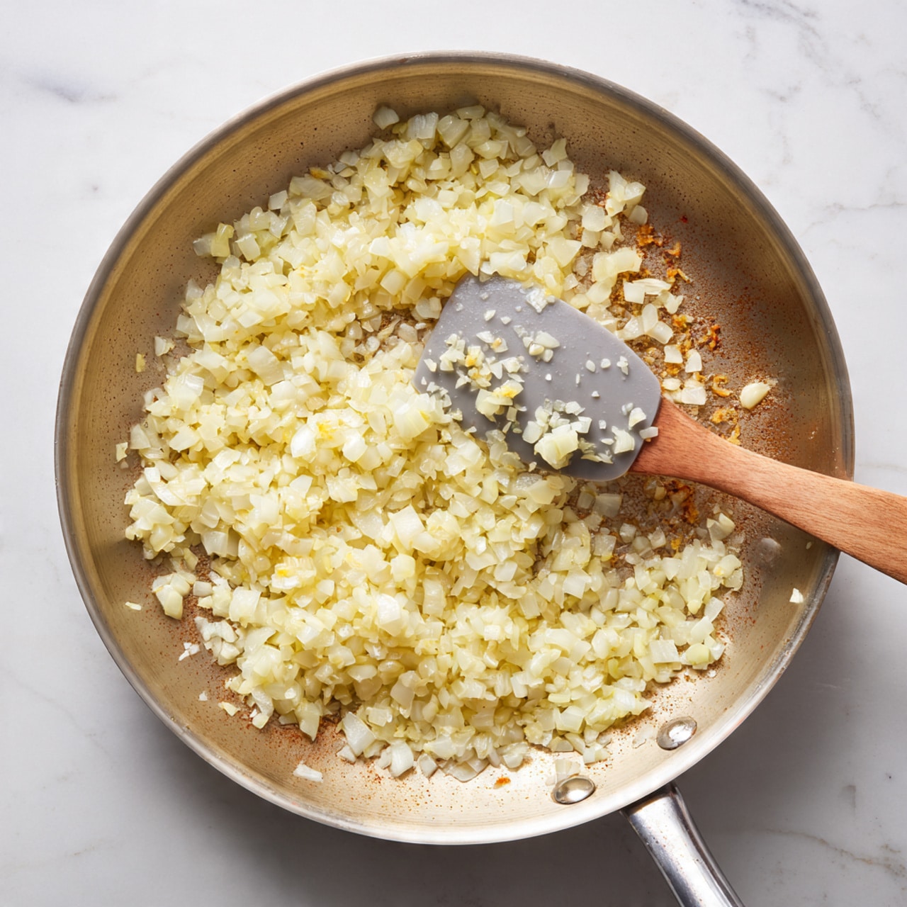 Inside a silver pan on a white marbled surface, there is a single layer of small chopped onions being cooked. The onions are light golden yellow with some pieces showing a slightly translucent texture, indicating they are softening as they cook. A gray silicone spatula is in the pan, stirring the onions, with small bits stuck to it. The pan has some brown bits on the bottom where the onions are cooking, adding a textured contrast to the smooth metal. photo taken with an iphone --ar 4:5 --v 7