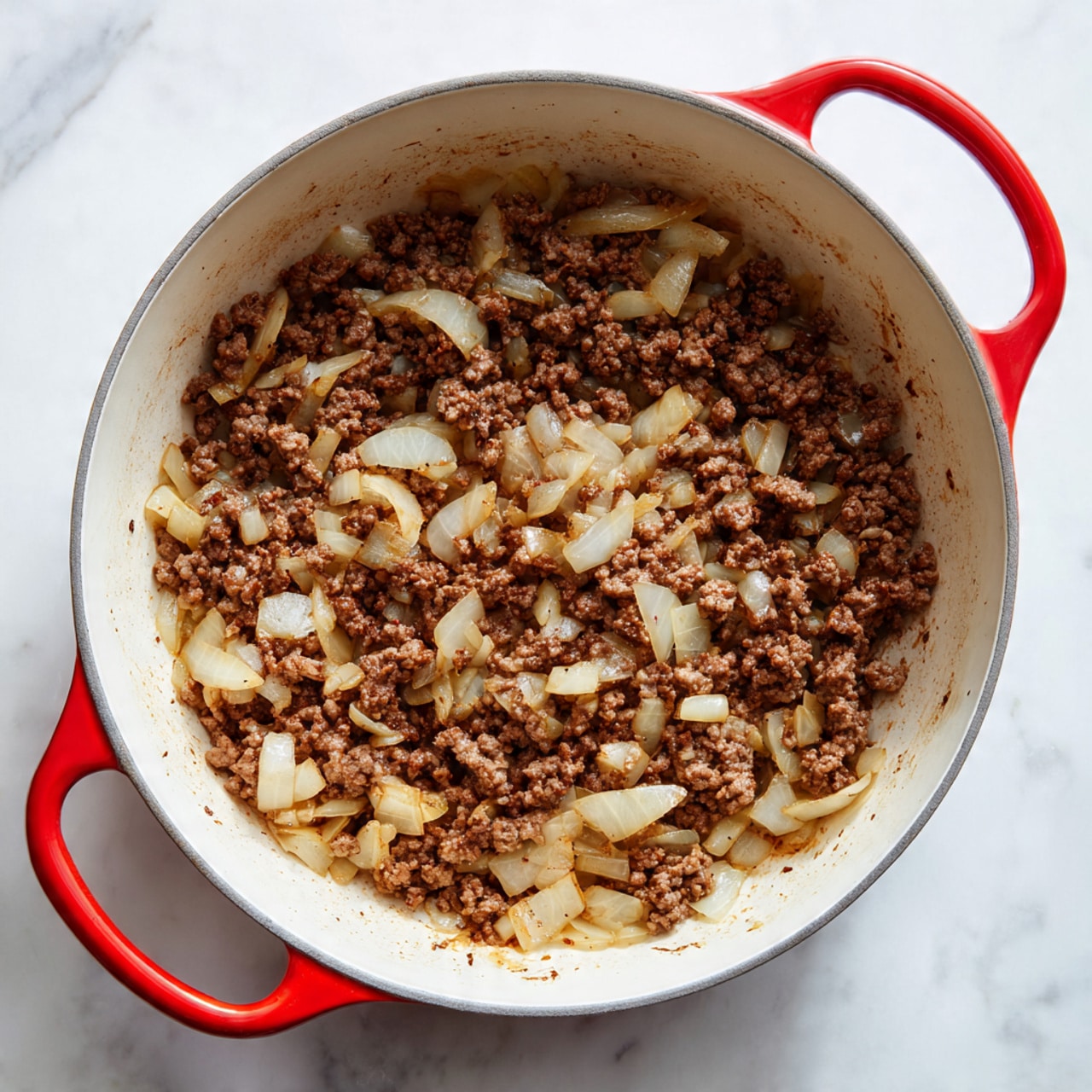 Inside a white pot with red handles, there is a single layer of cooked ground meat mixed with light translucent chunks of cooked onions. The meat is brown and crumbly, spread evenly across the bottom. The onions are soft and scattered throughout the meat, giving a mix of light and darker brown tones. The pot rests on a white marbled surface. photo taken with an iphone --ar 4:5 --v 7