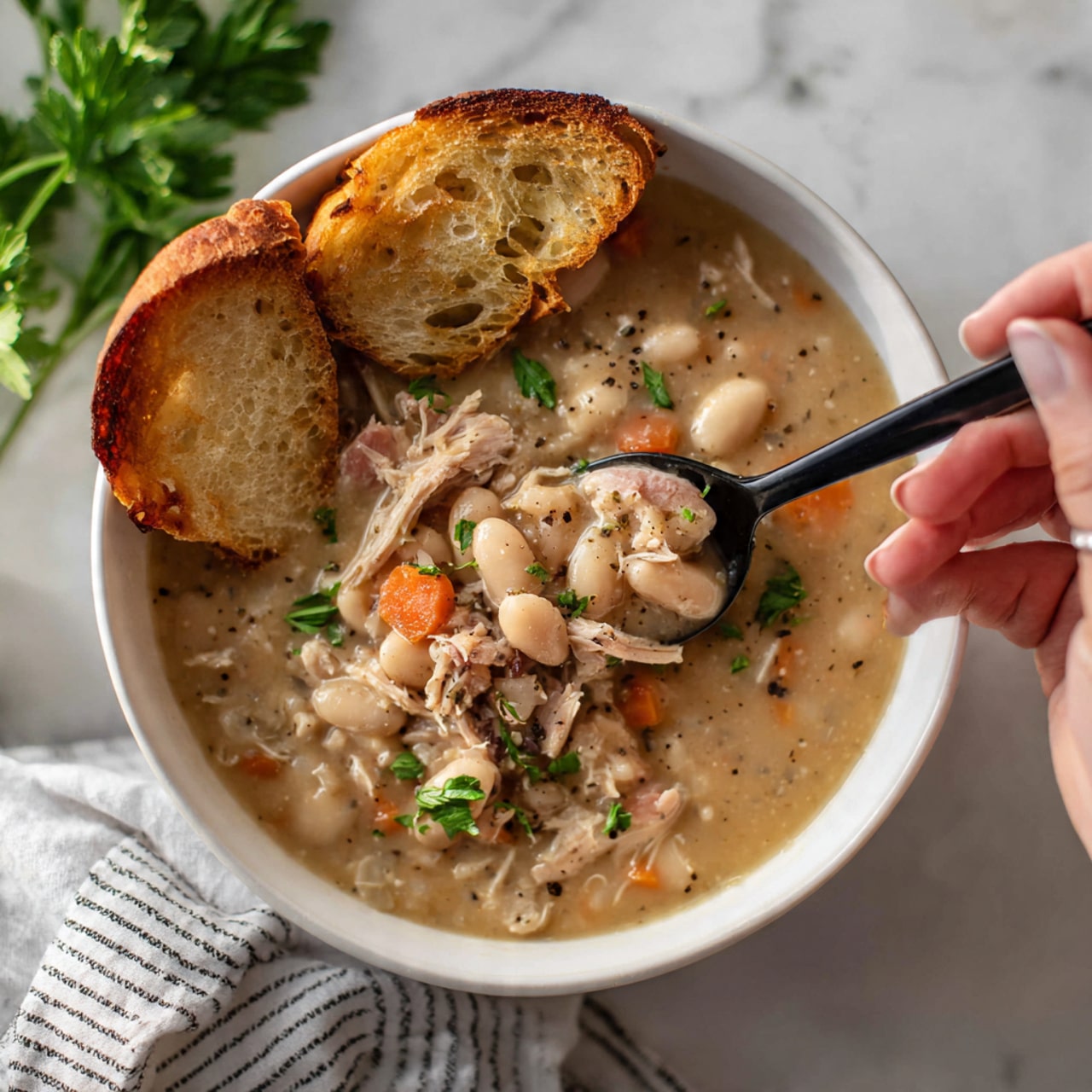 A white bowl filled with thick, creamy stew that has a light brown color speckled with small chunks of white beans, orange carrot pieces, and shredded pinkish-brown meat, all mixed with green parsley bits and black pepper. Two toasted slices of golden brown bread rest on the left edge of the bowl partly dipping into the stew. A black spoon is scooping some stew from the right side of the bowl, held by a woman's hand with part of a white and black striped cloth visible underneath. The bowl sits on a white marbled surface with fresh green parsley nearby. Photo taken with an iphone --ar 4:5 --v 7