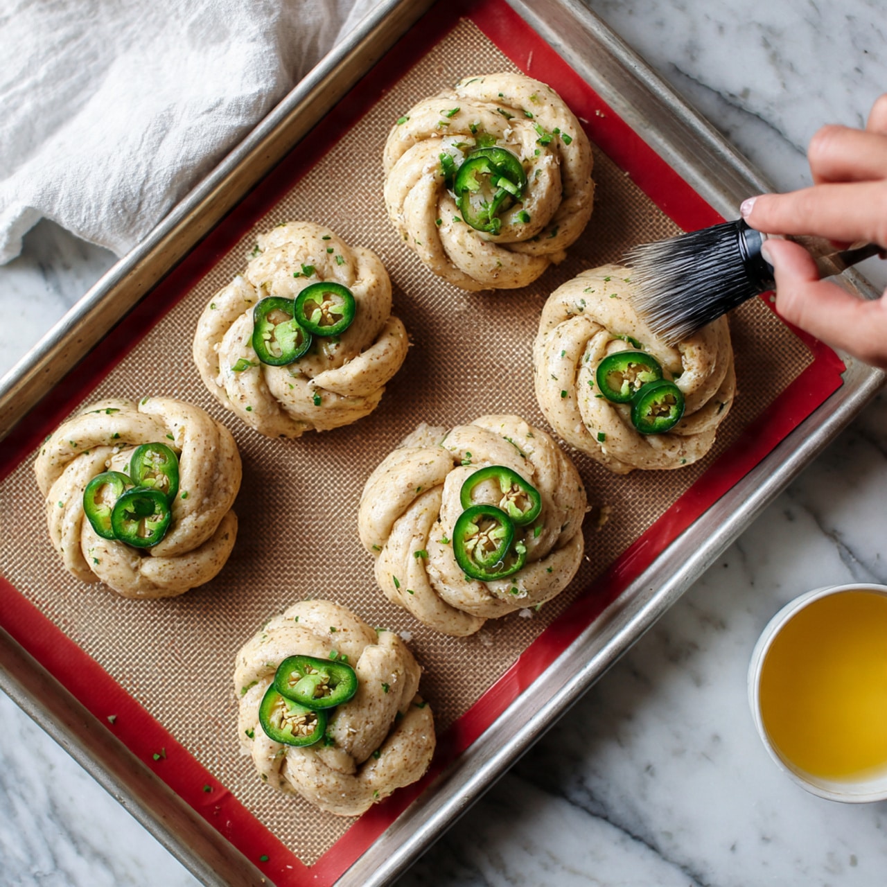 The image shows six raw pretzels on a light brown silicone mat placed on a silver metal baking tray with a red border. Each pretzel has a pale beige dough base twisted into a traditional pretzel shape. On top of the dough, there are three bright green jalapeño slices arranged on each pretzel, showing the seeds inside the jalapeño rounds. A woman's hand holds a black brush with light brown bristles, brushing the dough on the bottom right pretzel. The background is a white marbled surface, with a cup containing a yellow liquid placed on the right side. Photo taken with an iphone --ar 4:5 --v 7