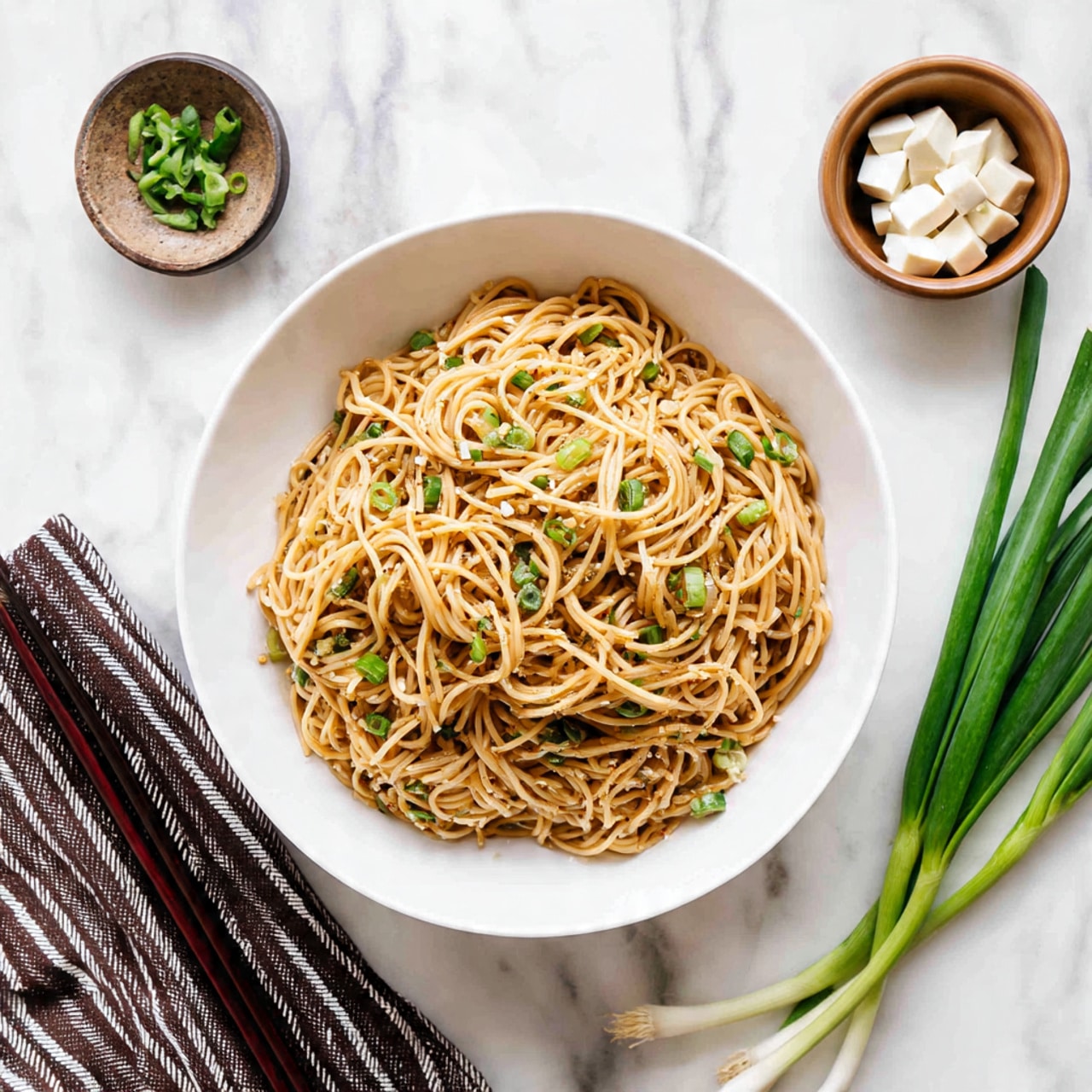 A white bowl filled with thin noodles tossed in a light brown sauce, with small pieces of chopped green onions mixed throughout, giving spots of fresh green color on the noodles. Surrounding the bowl on a white marbled surface are long green onions on the right side, a small brown bowl with white tofu pieces at the top right, and a second bowl of noodles partially visible at the top left. A striped cloth with dark brown chopsticks lies folded at the bottom left corner. The overall image is bright and clean with the white bowl and marbled background enhancing the warm tones of the noodles. photo taken with an iphone --ar 4:5 --v 7
