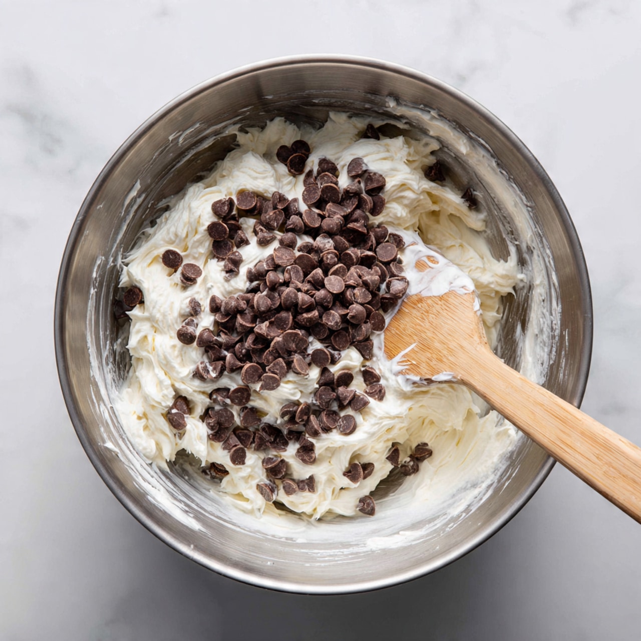 A shiny silver mixing bowl sits on a white marbled surface, filled with a thick, white creamy mixture. Dark brown chocolate chips are scattered evenly on top and partially mixed in, creating a textured look. A light wooden spatula rests in the bowl, with some of the white mixture clinging to it. The overall image shows a close view of the mixing process of a sweet treat photo taken with an iphone --ar 4:5 --v 7