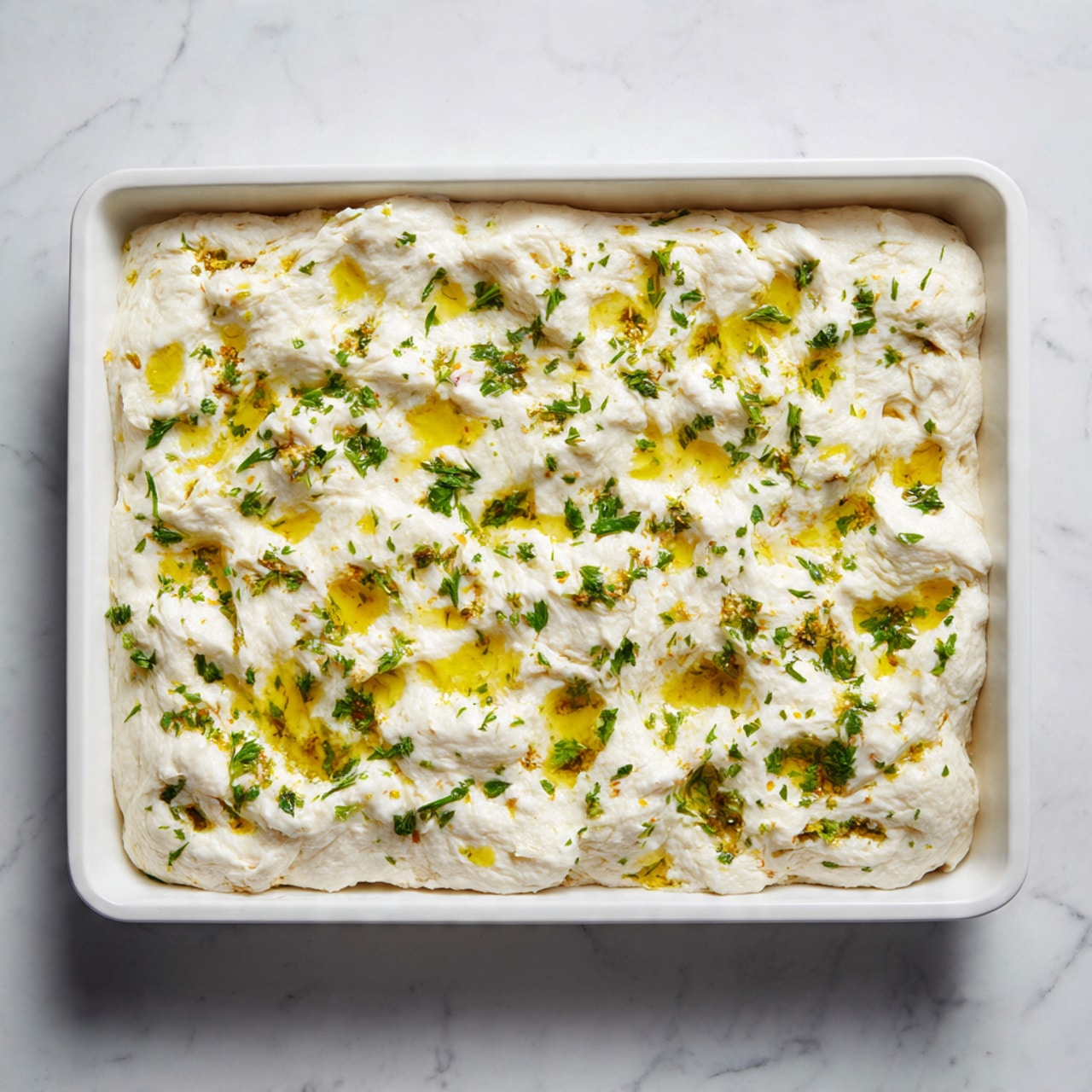 A rectangular white baking tray filled with a thick layer of light, fluffy dough that has a slightly uneven, puffy texture. The dough is dotted with small pools of golden yellow oil and sprinkled evenly with finely chopped green herbs scattered across the surface. The tray is placed on a white marbled background, highlighting the creamy white dough and the contrast of the green herb pieces and yellow oil. photo taken with an iphone --ar 4:5 --v 7