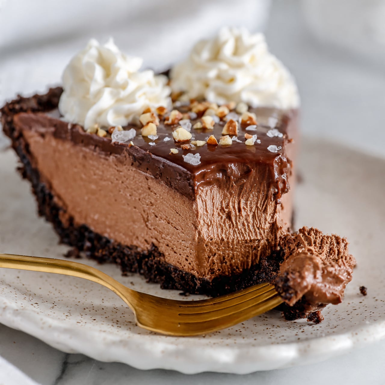 A slice of chocolate mousse pie on a white speckled plate with a scalloped edge, placed on a white marbled surface. The pie has three clear layers: the bottom layer is a dark, crumbly crust; the middle layer is a thick, smooth chocolate mousse with a light brown color and creamy texture; the top layer is a shiny dark chocolate ganache sprinkled with chopped nuts and small flakes of sea salt. Two swirls of white whipped cream sit on top near the back edge of the slice. In front of the pie slice, a gold fork holds a bite-sized piece showing the rich mousse texture and crust. Photo taken with an iphone --ar 4:5 --v 7