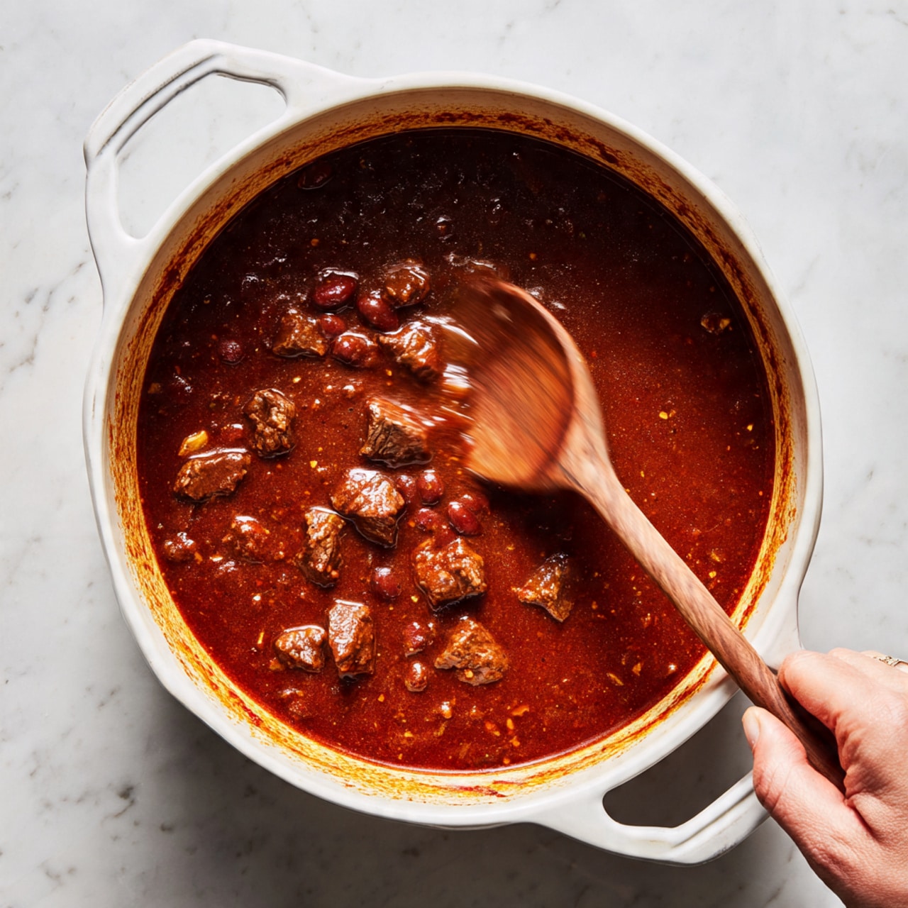 A white pot filled with thick, dark red stew that has visible chunks of browned meat and beans, showing a rich, textured surface. The stew is being stirred by a wooden spoon held by a woman's hand, creating a motion blur on the spoon. The pot has some reddish stains around the inside edge. The pot is placed on a white marbled surface with soft shadows. photo taken with an iphone --ar 4:5 --v 7