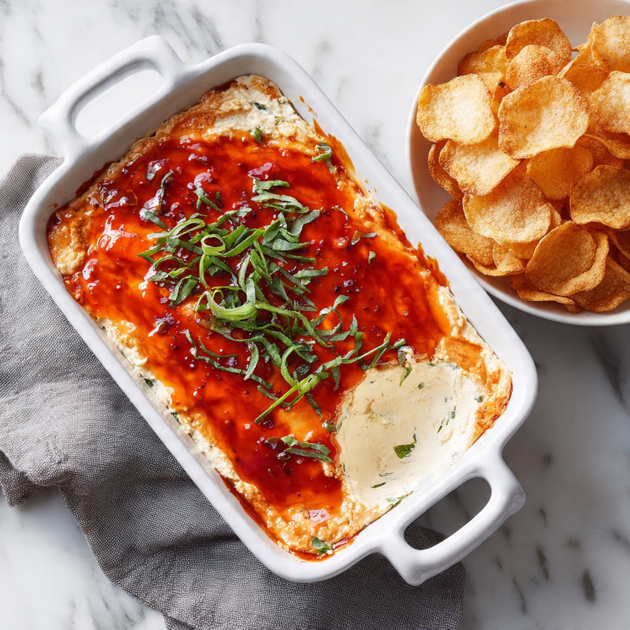 A white rectangular baking dish with two handles contains a creamy layered dip. The bottom layer is soft and pale, with a mix of melted cheese and creamy texture. The top layer is bright orange with a shiny red glaze spread evenly, and chopped fresh green herbs scattered on top, adding a fresh touch. Part of the dip has been scooped out from one corner, showing the creamy inside. Next to the dish, there is a white bowl filled with crispy, golden brown chips that have a bubbled texture. The items are placed on a white marbled surface with a folded gray cloth under the baking dish. photo taken with an iphone --ar 4:5 --v 7