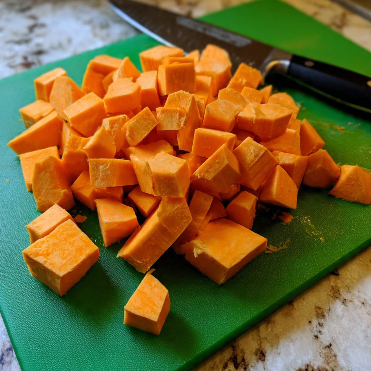 The image shows many pieces of orange sweet potato cut into chunks spread across a green cutting board. The pieces vary in size but mostly have a rough cube shape with smooth edges. Next to the sweet potato pieces, a large kitchen knife with a black handle lies flat on the cutting board. The background is a white marbled texture. photo taken with an iphone --ar 4:5 --v 7