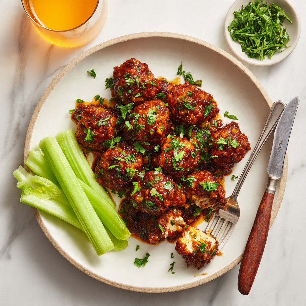 A white plate filled with ten pieces of fried chicken bites covered in a thick, dark reddish-brown sauce, garnished with small green herbs, arranged mostly in the center of the plate. On the left side of the plate, three bright green celery sticks lie parallel to the chicken. The plate itself is on a light beige bowl, placed on a white marbled surface. On the right edge of the plate, a silver fork with a wooden handle holds one chicken bite that is broken open, showing white inside meat. Above the plate, a small white bowl with chopped green herbs is partially visible, and a glass with orange liquid is on the left side of the image. Photo taken with an iphone --ar 4:5 --v 7
