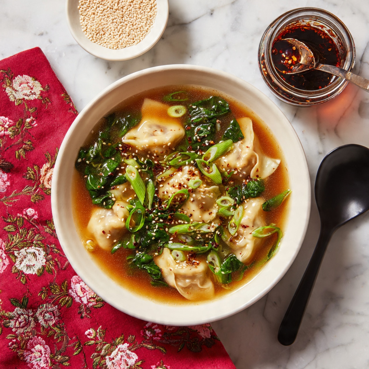 A white plate holds a dish of light brown dumplings in a clear brown broth. There are six dumplings arranged in the broth, with visible fresh green spinach leaves mixed throughout. The broth is thin and shiny with some red chili flakes floating on the surface. In the foreground, a black spoon holds one dumpling topped with a spinach leaf and some red chili flakes, highlighting the soft texture and folded edges of the dumpling. The scene is set on a white marbled surface. photo taken with an iphone --ar 4:5 --v 7