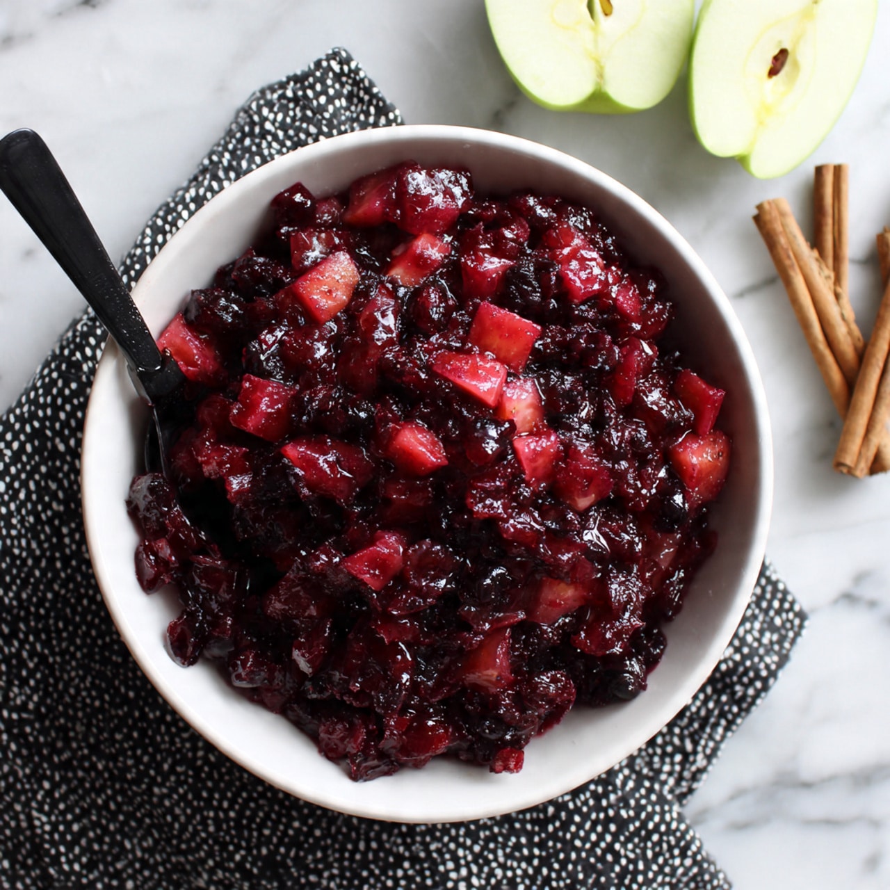 A white bowl filled with a thick mixture of dark red and bright red fruit pieces, mostly small chunks and bits that look soft and juicy, with a shiny texture. The mixture is heaped in the center of the bowl, and a black spoon is partially buried in it, with the spoon handle resting on the edge. The bowl sits on a black and white patterned cloth, which is placed on a white marbled surface. To the top right of the bowl, there are two halves of a pale green apple and three small cinnamon sticks slightly scattered nearby. Photo taken with an iphone --ar 4:5 --v 7