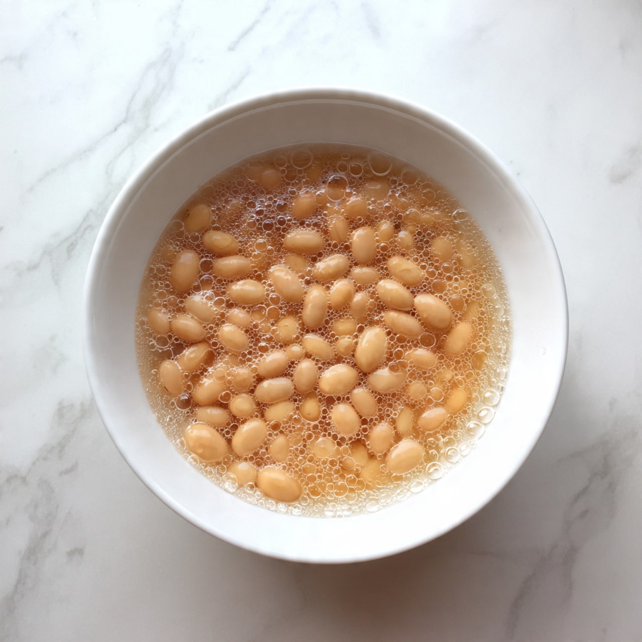 A white bowl filled with a layer of small beige beans soaking in clear water, with some small bubbles floating on the surface. The bowl sits on a white marbled surface, giving a clean and fresh look. The beans are evenly spread and partially submerged, showing smooth and rounded shapes. photo taken with an iphone --ar 4:5 --v 7