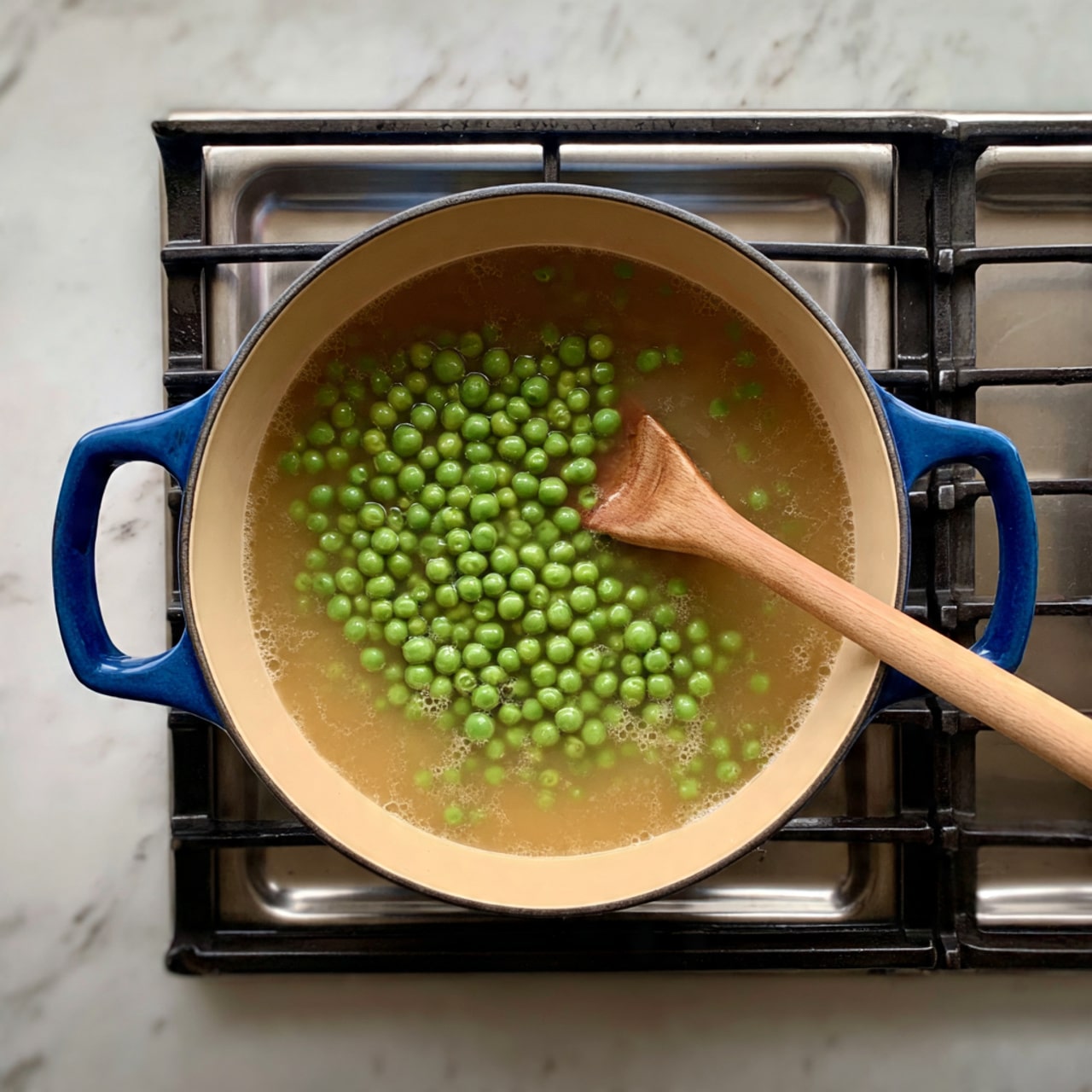 A blue cooking pot with a beige inside sits on a stove with black grates, filled with a light brown broth containing many small green peas floating on top, mixed gently with a wooden spoon resting inside the pot. The stove is next to a white marbled surface. Photo taken with an iphone --ar 4:5 --v 7