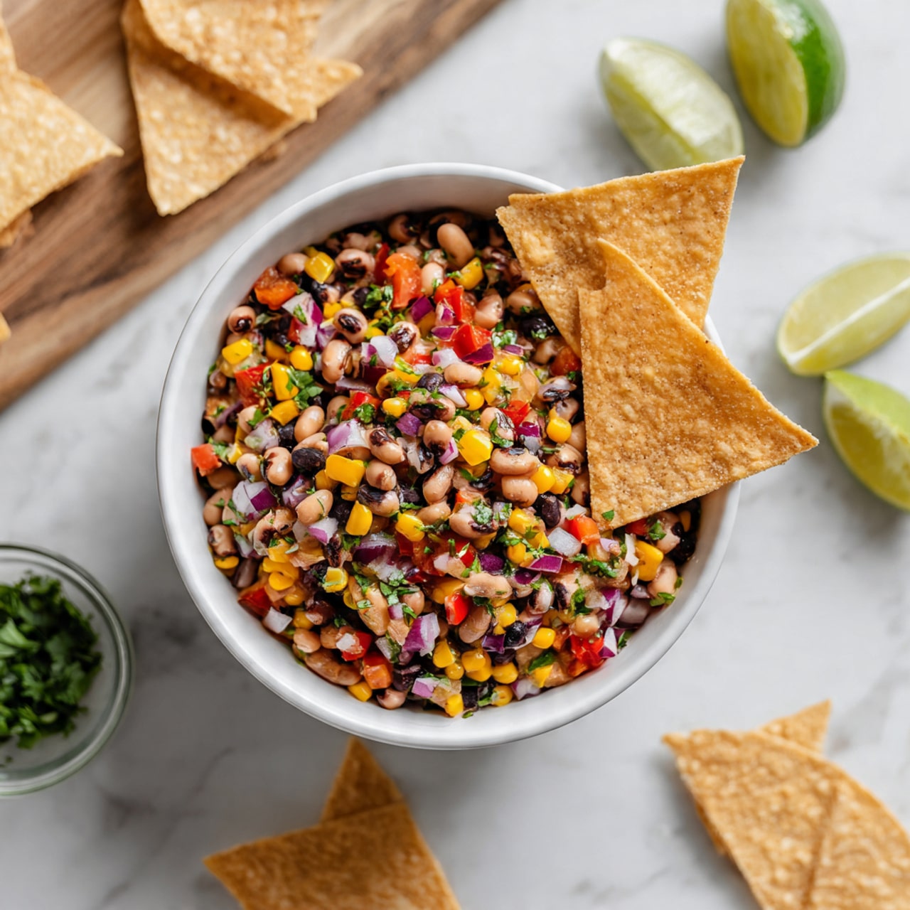 A white bowl filled with a colorful salad consisting of layers of black-eyed peas, black beans, yellow corn kernels, diced red bell peppers, finely chopped red onions, and small pieces of green herbs mixed evenly throughout. Three light beige, round-edged tortilla chips are placed upright inside the salad near the right side of the bowl. The bowl sits on a white marbled surface surrounded by scattered tortilla chips and a wooden cutting board with lime wedges, one lime cut in half showing its juicy, pale green inside. A clear glass bowl with chopped cilantro is partially visible on the left side. The scene is bright and fresh, emphasizing the textures and vibrant colors of the salad ingredients photo taken with an iphone --ar 4:5 --v 7