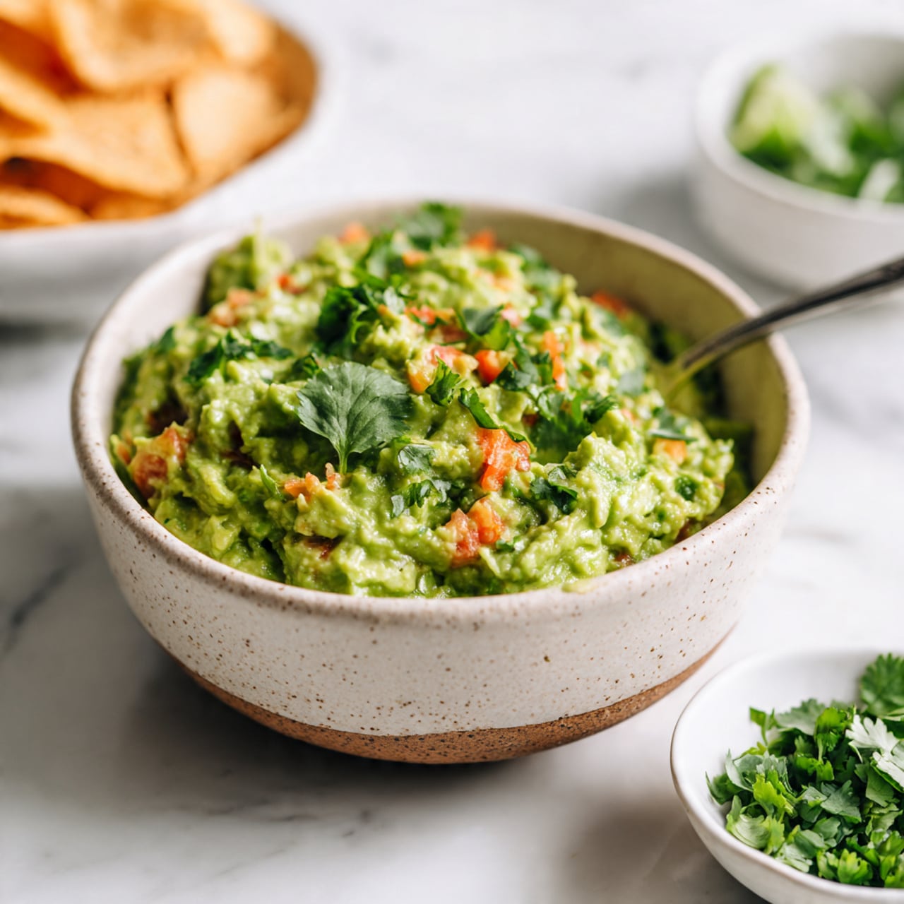 The image shows a bowl of guacamole with a rough, chunky texture featuring bright green mashed avocado mixed with small pieces of red tomato and green cilantro leaves, giving it a colorful and fresh look. The bowl is beige with a speckled pattern, filled to the top with the guacamole, and has a silver spoon sticking into it. Nearby, there is a small white bowl containing chopped cilantro and a white bowl partially visible with green vegetables inside. To the left, tortilla chips rest in a white bowl, showing a light, crispy texture and golden color. The whole set is placed on a white marbled surface. photo taken with an iphone --ar 4:5 --v 7
