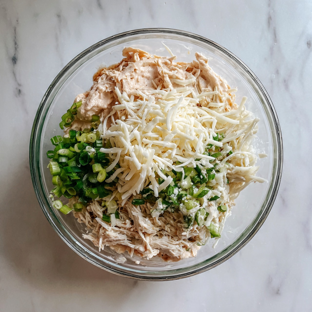 A clear glass bowl sits on a white marbled surface filled with three separate layers of ingredients: one side holds a creamy light pink sauce, the next side has white shredded cheese, and the last side shows chopped green onions with a bit of white shredded chicken on top. The layers are unmixed and clearly visible in the bowl. photo taken with an iphone --ar 4:5 --v 7