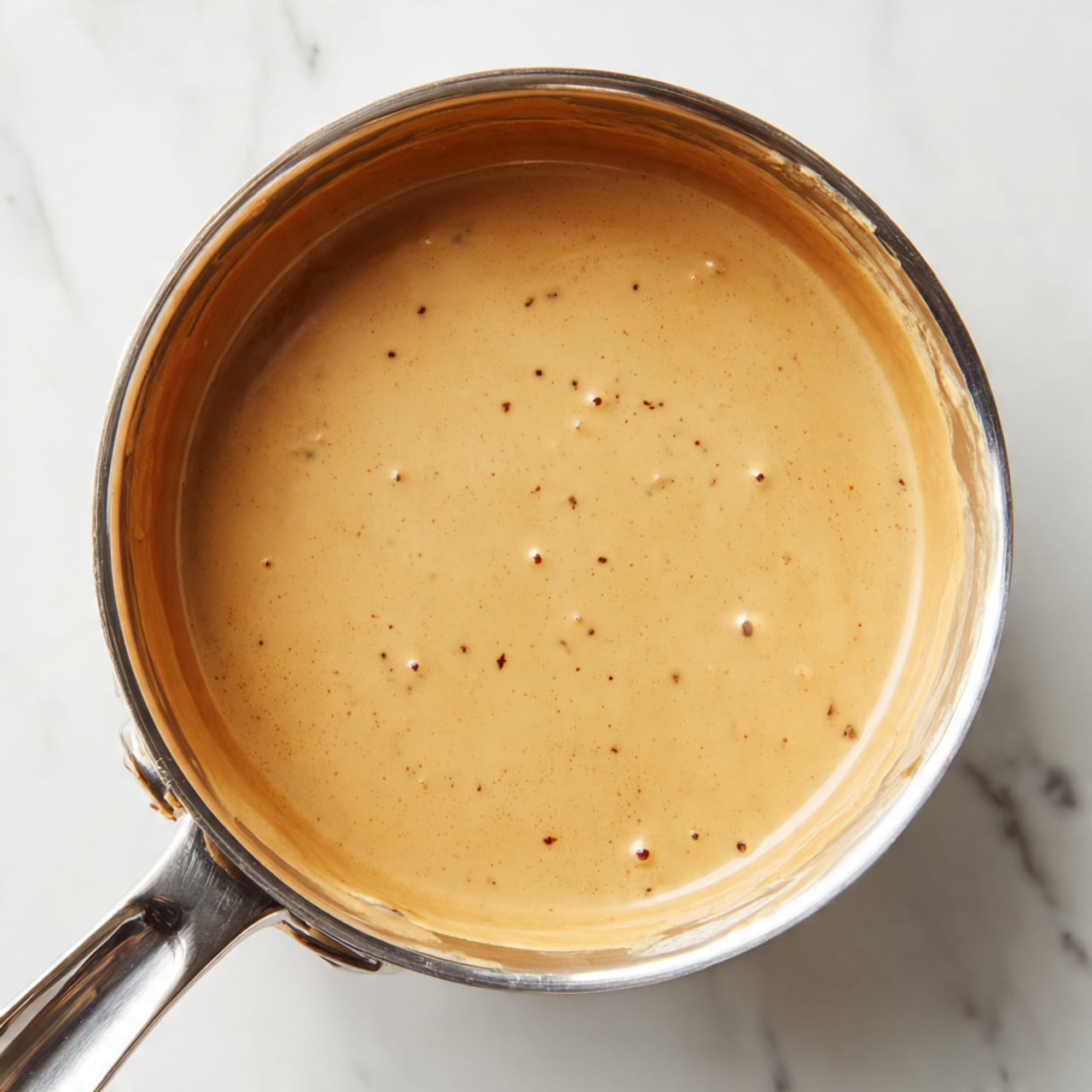 A top view of a metal saucepan filled with a smooth, creamy light brown sauce. The sauce has a few small bubbles and tiny dark specks scattered on the surface. The inside walls of the pan show slight streaks of the sauce clinging to them. The saucepan handle is shiny and metallic, extending out towards the bottom left corner. The pan is placed on a white marbled surface. photo taken with an iphone --ar 4:5 --v 7