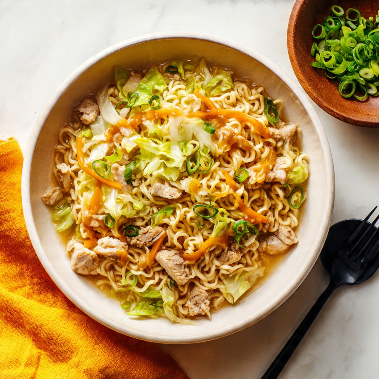 A large silver pan filled with a noodle dish showing three main layers: at the bottom, light brown cooked chicken pieces scattered unevenly; the middle layer has pale beige cooked ramen noodles mixed with translucent, wilted cabbage; the top layer features thin strips of bright orange carrots and chopped green onions sprinkled all over. A black spatula with a wooden handle rests inside the pan, positioned on the right side. The pan is placed on a white marbled surface with a yellow cloth underneath its handle, a wooden bowl of green onions, and pieces of ginger near the top right. Photo taken with an iphone --ar 4:5 --v 7