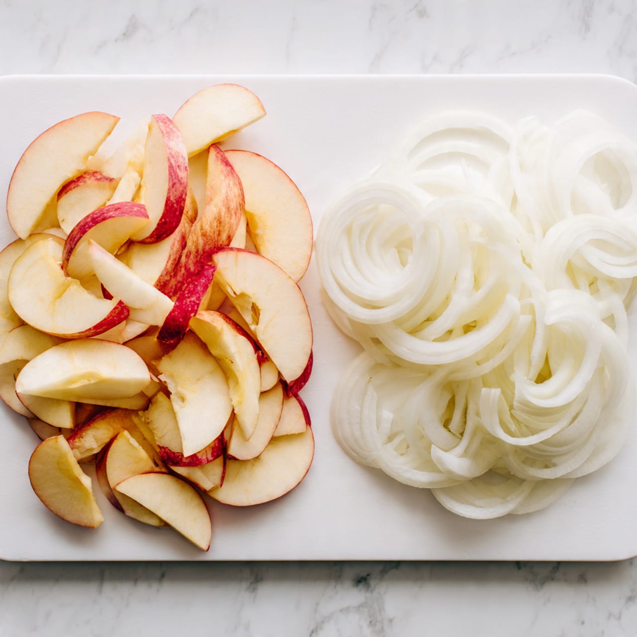 A white cutting board sits on a white marbled surface, holding two piles of sliced ingredients side by side. On the left, there are many light yellow apple slices with red skin edges, arranged in a loose pile. On the right, there are thinly sliced white onion rings, layered loosely in a similar pile. The colors show soft yellows, reds, and white with smooth textures on the apple and thin, slightly translucent layers on the onion. Photo taken with an iphone --ar 4:5 --v 7