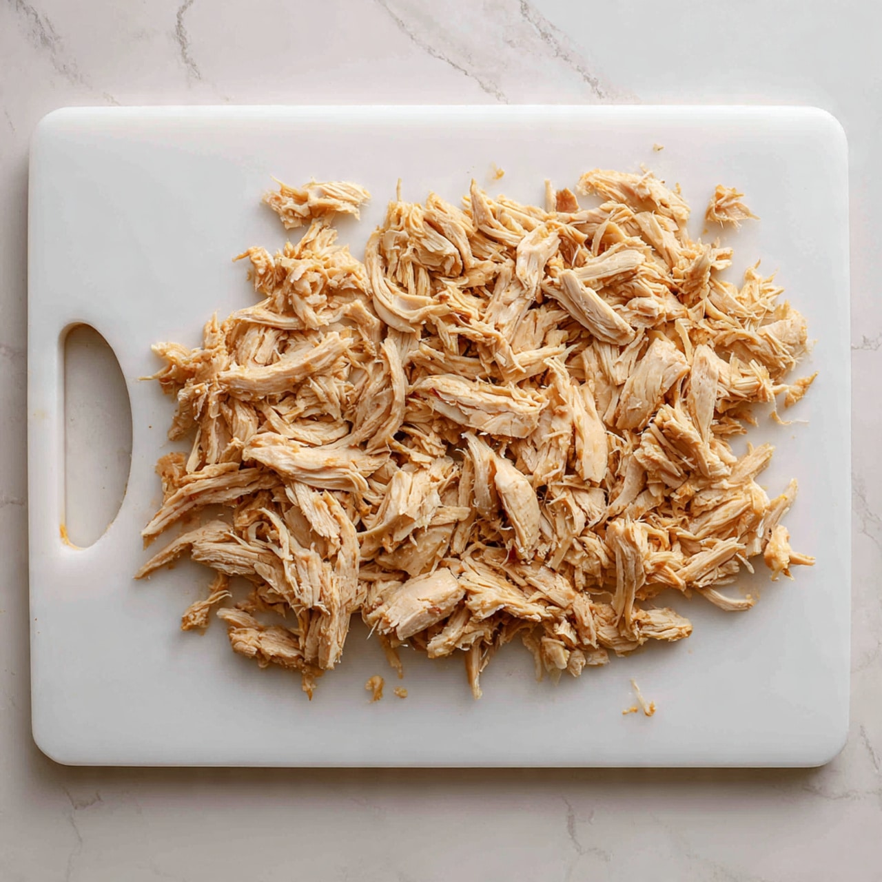 The image shows shredded cooked chicken spread evenly on a white cutting board placed on a white marbled surface. The chicken pieces vary in size and shape, mainly thin strands with some thicker pieces mixed in. The chicken is light brown with some parts slightly darker, showing texture and cooked surface. The board has a rounded corner with a handle on the left side. The background is clean and simple, focusing on the chicken on the board. Photo taken with an iphone --ar 4:5 --v 7