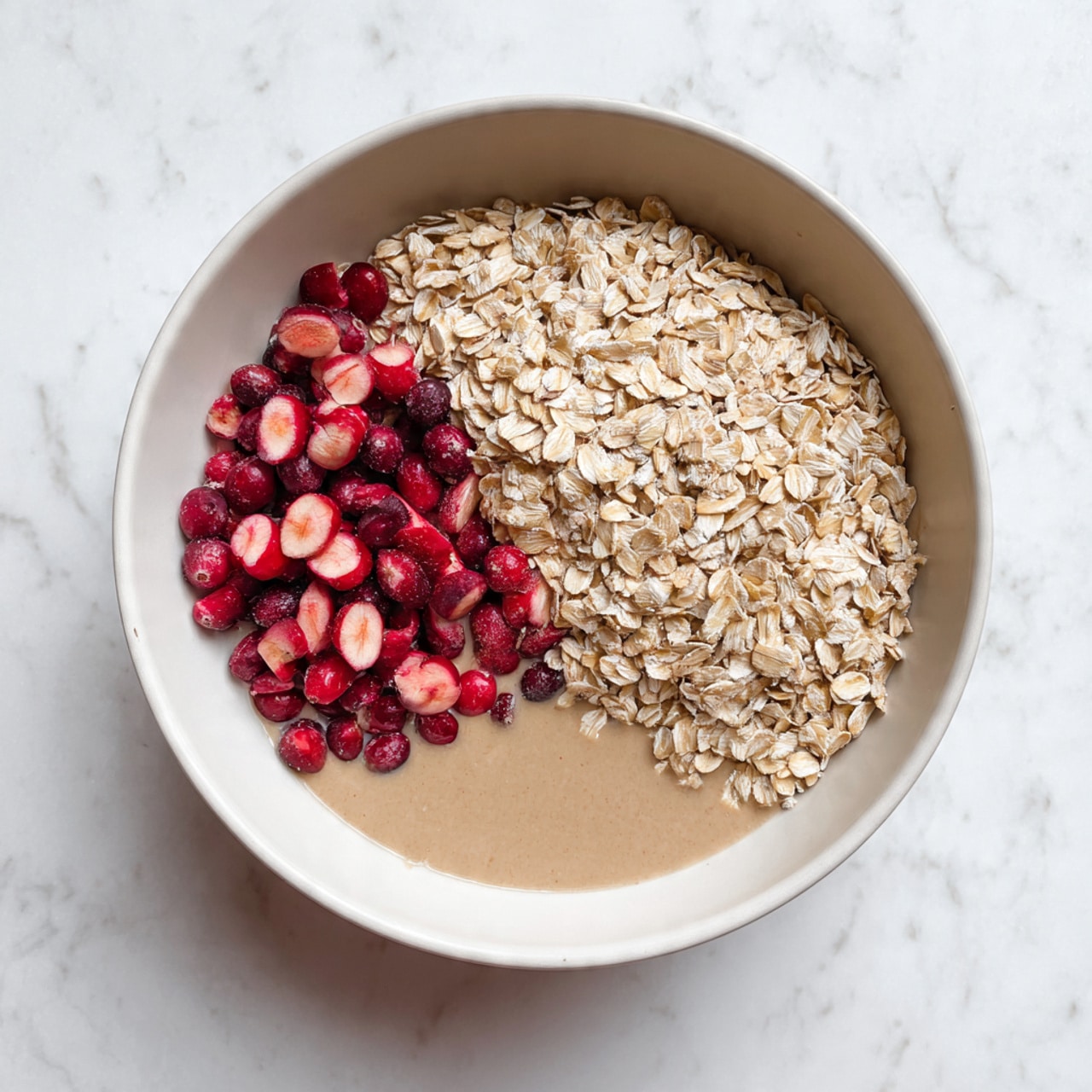 A large white bowl sits on a white marbled surface, filled with three main layers of ingredients. The first layer at the bottom is a light brown, smooth liquid mixture. On the right side, covering almost half of the bowl, there is a thick layer of dry, pale beige rolled oats with a rough texture. On the left bottom side, there is a cluster of fresh cranberries, some whole and some sliced in half, showing a bright red outer skin and pale pink inside. Photo taken with an iphone --ar 4:5 --v 7