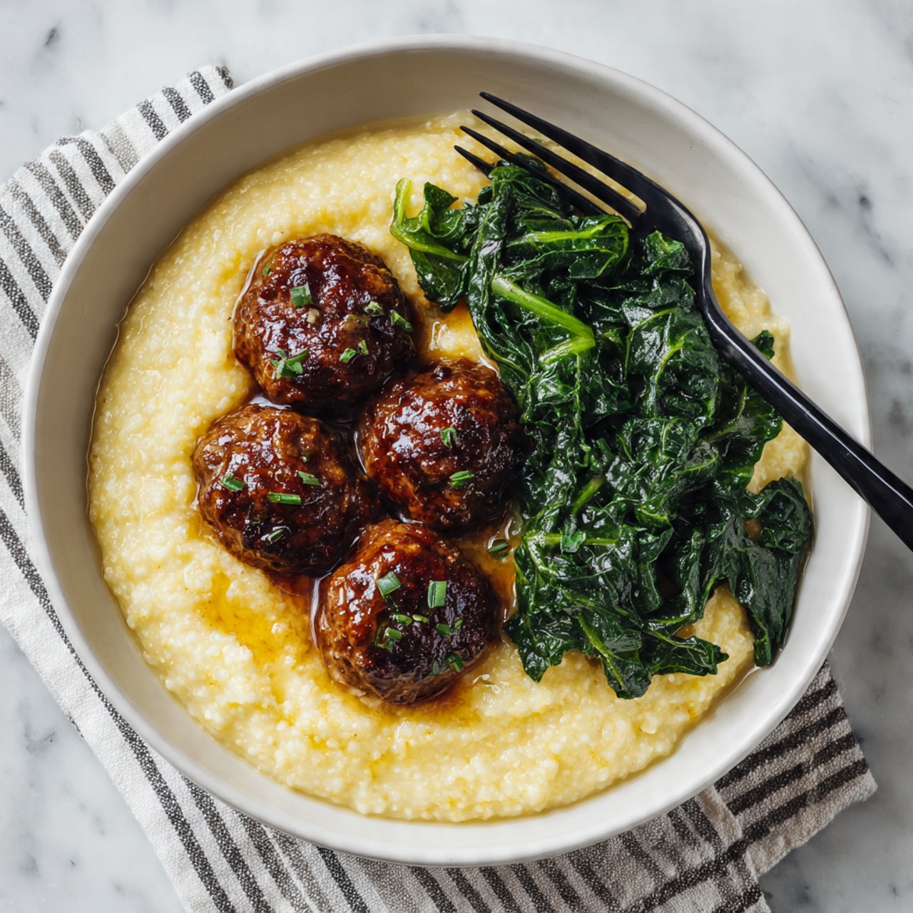The image shows a white bowl filled with three main layers: the bottom layer is creamy, pale yellow grits with a slightly grainy texture, topped with four glazed, shiny brown meatballs placed near the center. To the right side of the bowl is a serving of cooked dark green leafy vegetables with a soft, wilted texture. A black fork is resting on the edge of the bowl above the vegetables. The bowl is placed on a white marbled surface with a striped cloth partially visible. Photo taken with an iphone --ar 4:5 --v 7