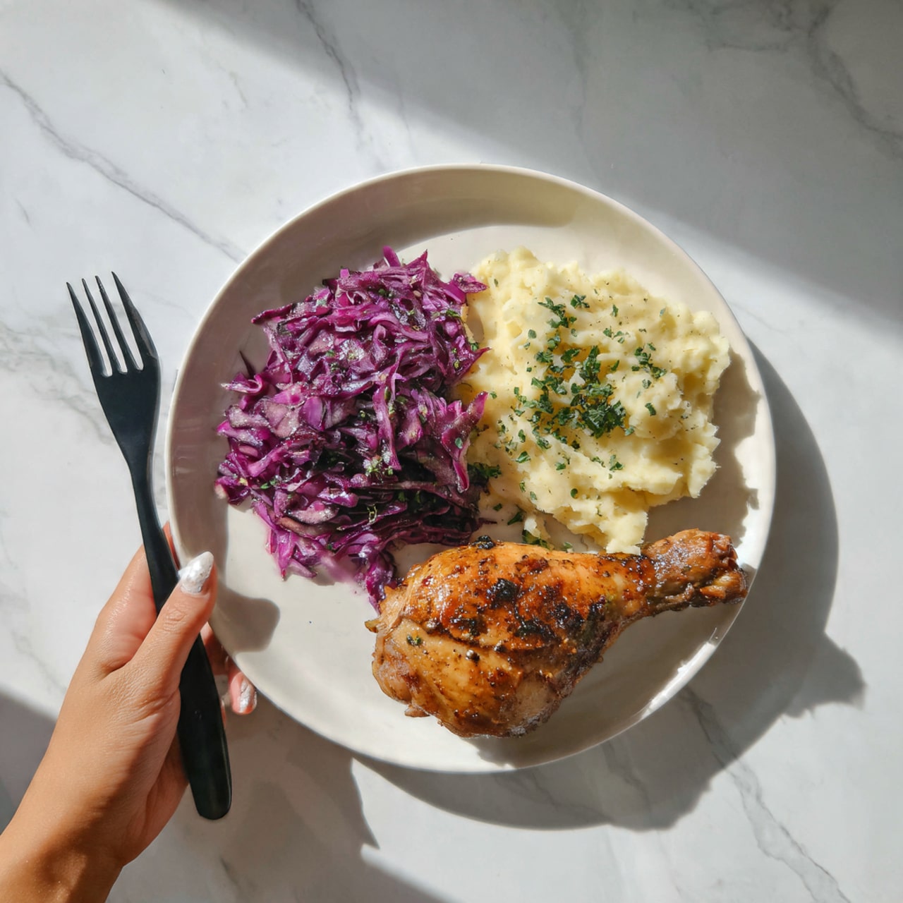 A white plate on a white marbled surface holds a meal with three layers: on the left side is bright purple cabbage with some green herb pieces mixed inside, next to it on the top right is a portion of creamy, light beige mashed potatoes sprinkled lightly with green herbs, and on the bottom right is a single cooked chicken leg with golden brown, slightly charred skin and small green herb bits on top. A black fork is placed to the left of the plate, and a woman's hand holds it gently. The lighting is natural and bright, showing details in colors and textures. photo taken with an iphone --ar 4:5 --v 7