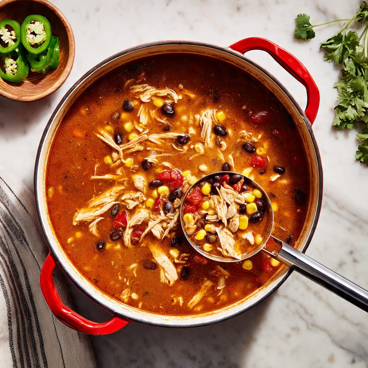 A large round white pot with red handles is filled with a thick reddish-brown soup containing shredded pieces of light brown chicken, yellow corn kernels, black beans, and diced red tomatoes. A metal ladle with a black handle scoops some of the soup, showing the mixture of ingredients clearly. To the left side, there is a small bowl with sliced green jalapeño peppers on a white marbled surface. A striped cloth is partially visible to the lower left of the pot, and some fresh green herbs are placed to the upper right on the white marbled surface. Photo taken with an iphone --ar 4:5 --v 7