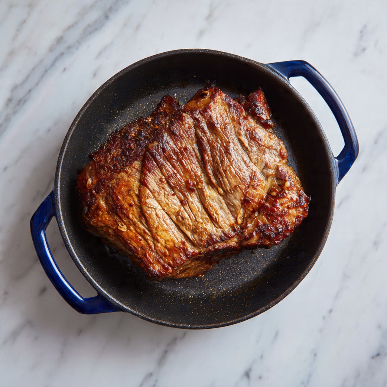 The image shows a large piece of cooked meat with a browned, slightly crispy surface, lying flat inside a black cast iron pan with two blue handles on each side. The meat has visible texture and light grill marks in some areas, with different shades of brown, from light to dark. The pan rests on a white marbled surface, which gives a clean and simple background. The overall look is rustic and warm, focusing on the meat and the cast iron pan. photo taken with an iphone --ar 4:5 --v 7