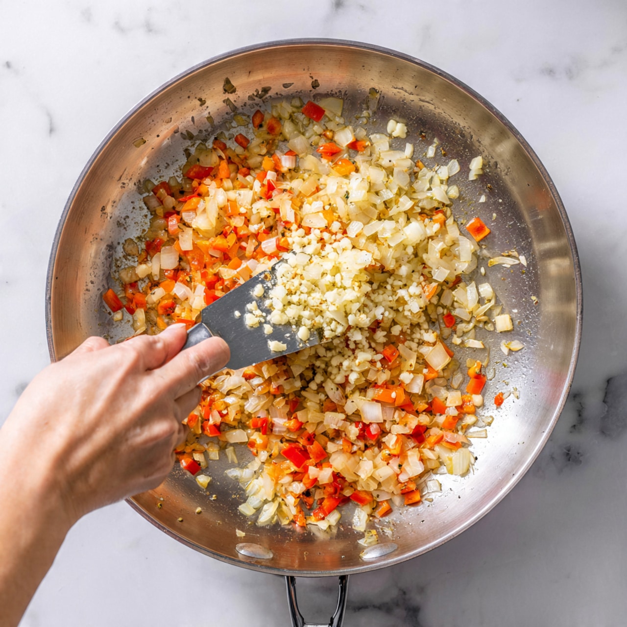 A close-up top view of a stainless steel pan sitting on a white marbled surface, with finely chopped onions and red bell peppers sautéing inside, showing soft and slightly translucent textures of the onions mixed with the bright orange-red bits of the peppers; a woman's hand is holding a metal scraper adding finely chopped garlic to the pan, the garlic pieces are pale and fresh-looking, creating a mix of three colorful layers inside the pan. Photo taken with an iphone --ar 4:5 --v 7