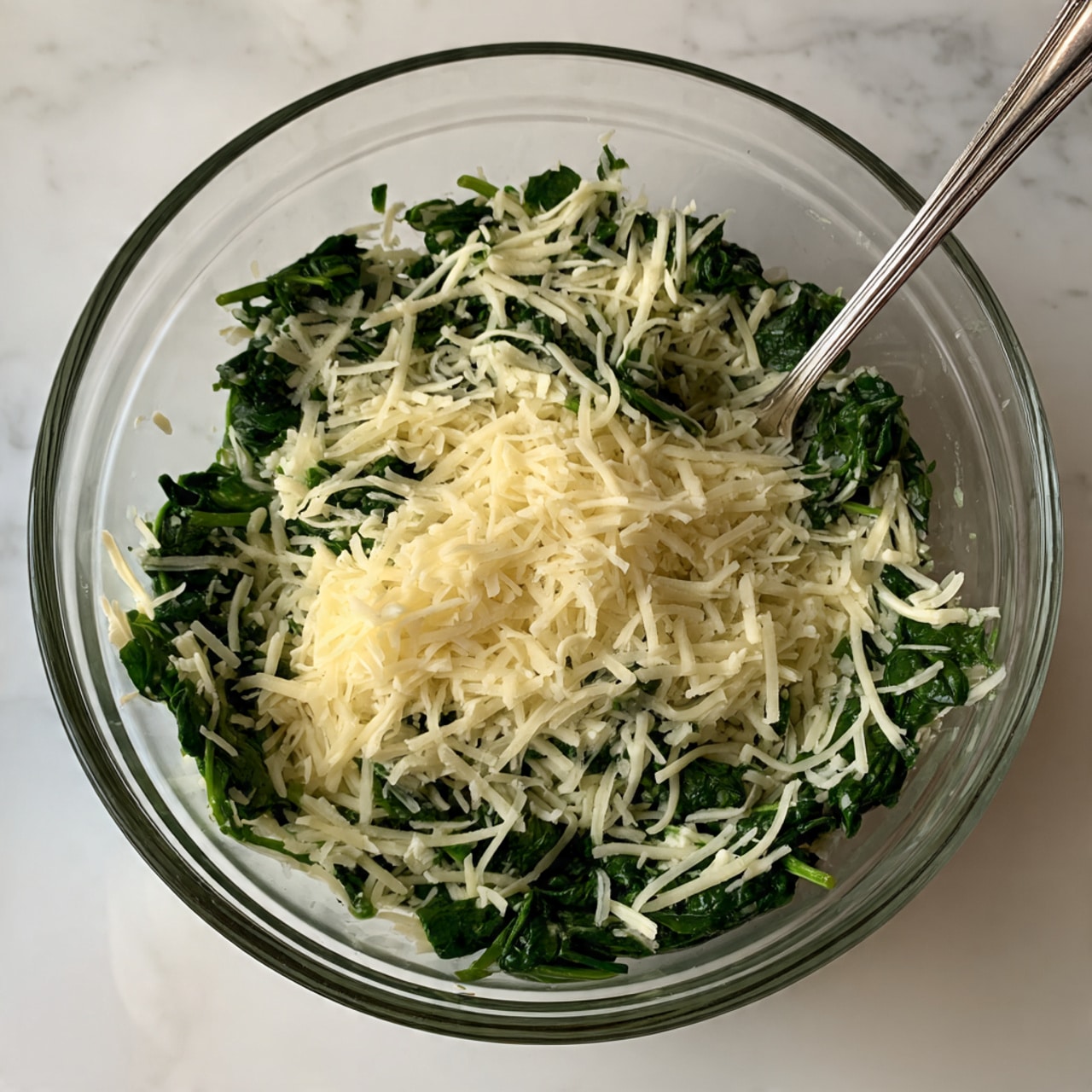 A clear glass bowl contains two layers mixed together: bright dark green chopped spinach and thin, white shredded cheese. The spinach pieces are small and leafy, spread evenly with the cheese strands scattered throughout. A silver fork rests inside the bowl on the right side, slightly touching the mix. The bowl is on a white marbled surface with soft lighting. Photo taken with an iphone --ar 4:5 --v 7