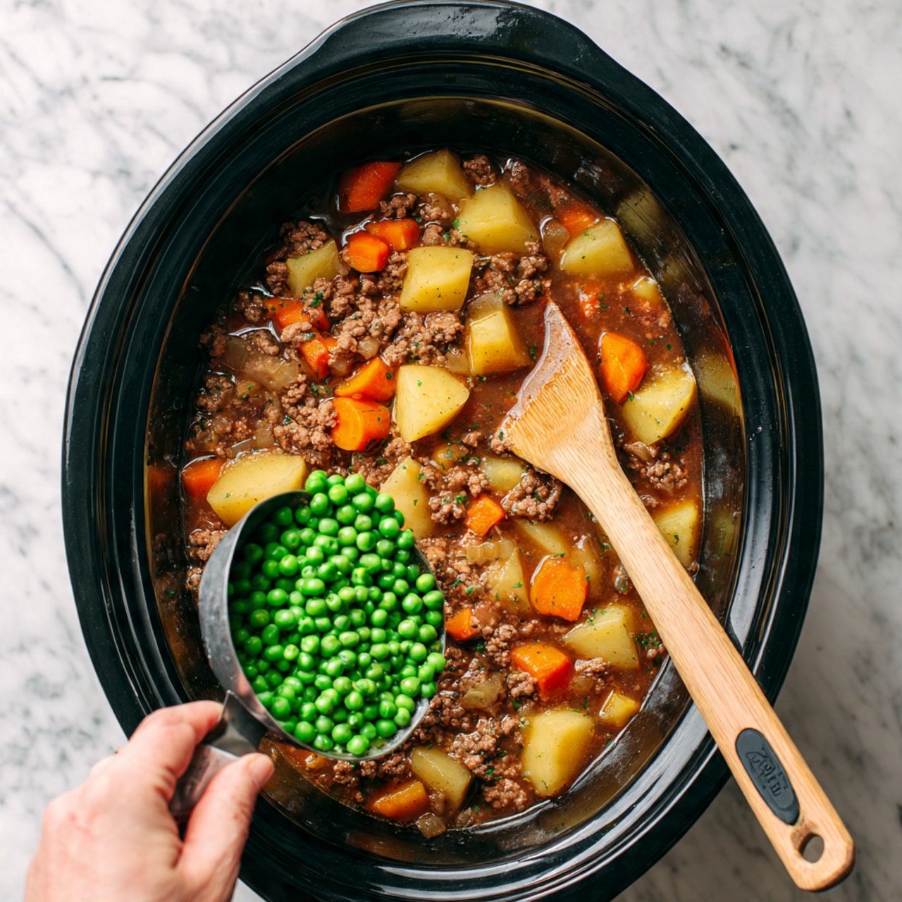 A black slow cooker is filled with a stew-like mixture showing chunks of light yellow potatoes, bits of orange carrots, small pieces of browned ground meat, and a dark brown liquid broth, all mixed together. A wooden spoon with a black silicone head rests in the cooker on the right side. From the left bottom corner, a woman's hand is pouring bright green peas from a metal measuring cup into the slow cooker. The scene is set on a white marbled surface. photo taken with an iphone --ar 4:5 --v 7
