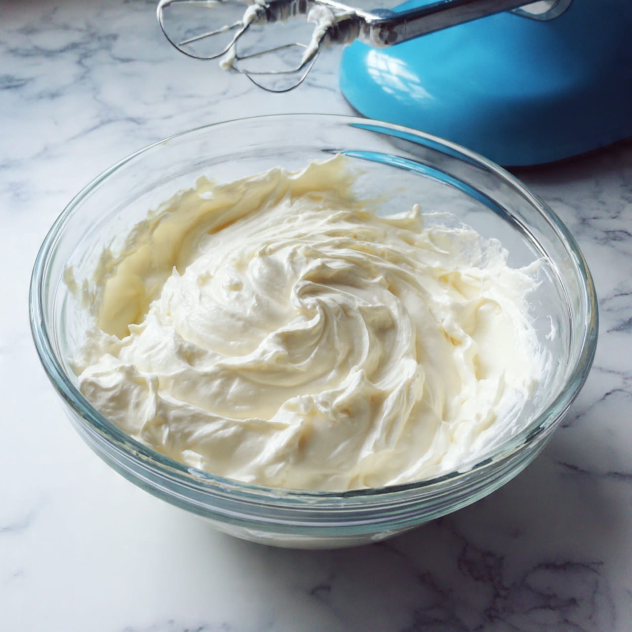 A woman's hand with black nail polish holds a metal whisk attachment covered with a smooth, thick layer of white whipped cream or meringue forming a peak at the top, with some cream trailing down the thin metal wires of the whisk. The background shows a soft, out-of-focus light gray texture. Photo taken with an iphone --ar 4:5 --v 7