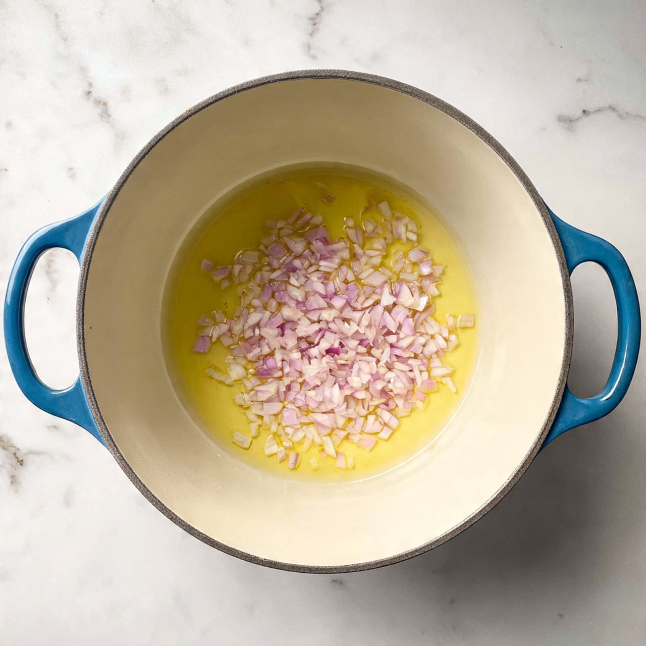 The image shows a white pot with two blue handles placed on a white marbled surface. Inside the pot, there is one visible layer of light yellow melted butter covering the bottom, topped with a layer of finely chopped shallots that are pale pink and white in color. The pot has a smooth, cream-colored interior. Photo taken with an iphone --ar 4:5 --v 7