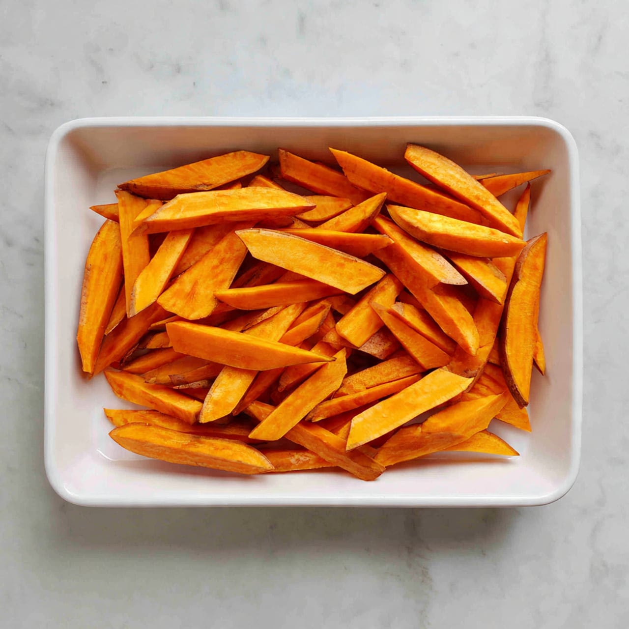 A white rectangular tray sits on a white marbled surface, filled with one layer of evenly spread sweet potato sticks. The sticks are bright orange with some parts showing light brown edges, thinly sliced into long, rectangular shapes with pointed tips, arranged mostly parallel but slightly overlapping. The tray has a clean, simple look with the sweet potatoes taking up most of the space, creating a neat and organized visual. photo taken with an iphone --ar 4:5 --v 7