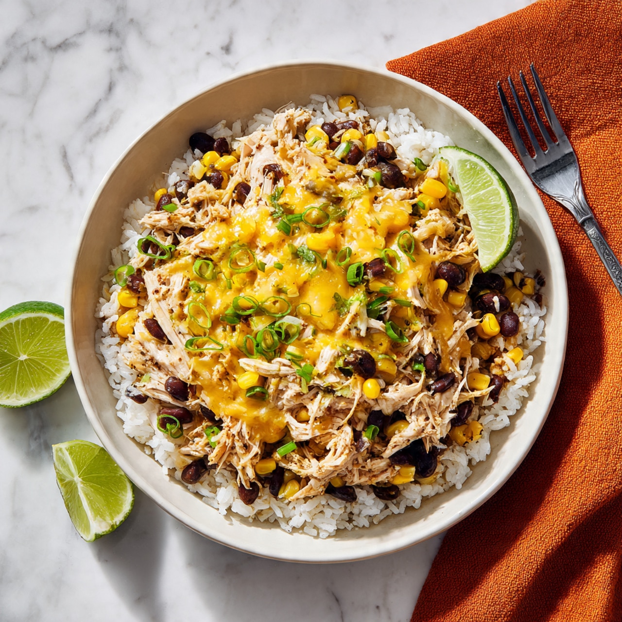 The image shows a white bowl filled with layers of food placed on a white marbled surface. The bottom layer is white rice, topped with shredded chicken mixed with black beans and yellow corn kernels. Melted yellow cheese covers the top with some green chopped herbs and green onions scattered around. There are two lime wedges tucked on one side of the bowl. A silver fork rests nearby on a folded orange cloth. The lighting is bright, making the colors vivid and fresh. photo taken with an iphone --ar 4:5 --v 7