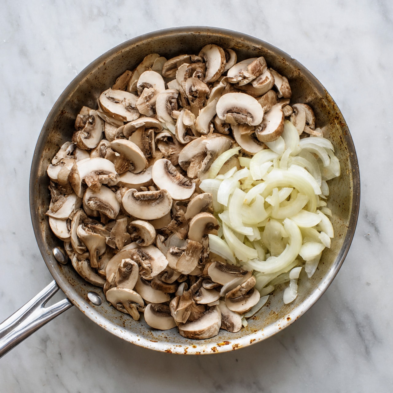 A silver pan filled with thinly sliced light brown mushrooms and white onion slices. The mushrooms cover most of the pan's bottom in a scattered pile, while the onions are grouped on one side. The pan is placed on a white marbled surface, showing some spots where the pan has browned the pan's bottom. The pan has two handles, a long one and a small round one. Photo taken with an iphone --ar 4:5 --v 7