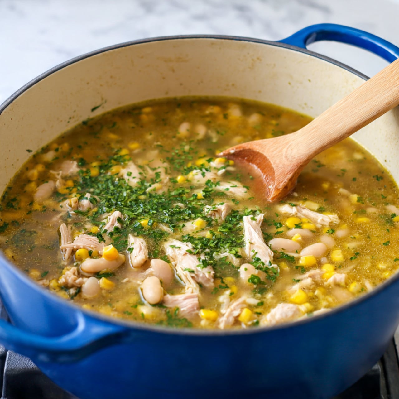 A large blue cooking pot filled with a warm soup sits on a stove. The soup has several layers: a clear, golden broth with small pieces of white beans and yellow corn spread throughout. There are chunks of light beige cooked chicken mixed in, topped with bright green chopped herbs. A wooden spoon is partially stirring the soup from the right side, touching the surface. The stove is black, and the background surface is white marbled. Photo taken with an iphone --ar 4:5 --v 7