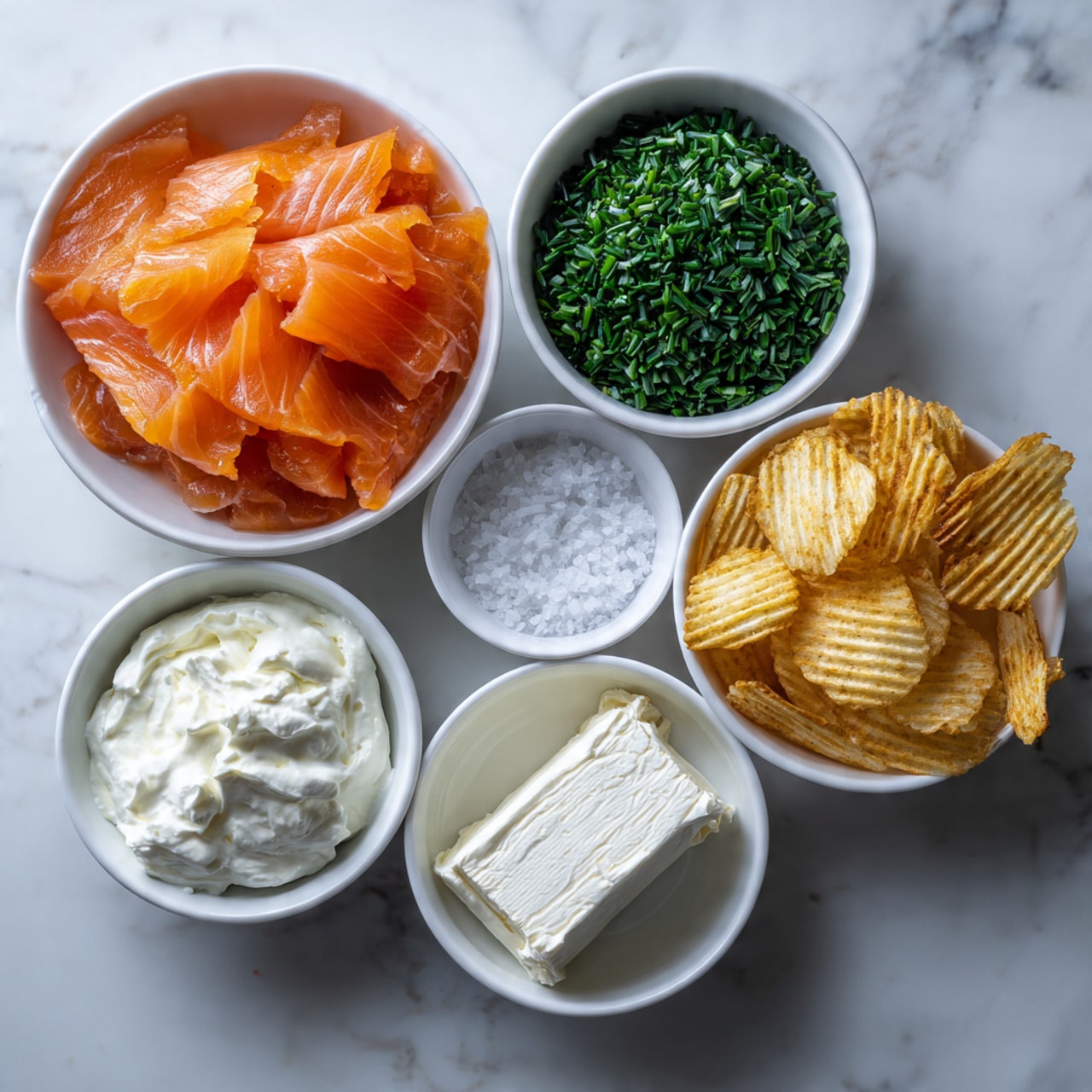 The image shows six white bowls on a white marbled surface, each with a different ingredient. The largest bowl on the left contains thin, layered slices of orange smoked salmon with a shiny, smooth texture. To the right of it, the top middle bowl holds bright green chopped chives with a fresh, leafy look. Below that is a small bowl with white sea salt crystals. The top right bowl is filled with golden ridged potato chips, stacked in a crinkly texture. The bottom left bowl contains dark green, round capers. The bottom middle bowl has thick, smooth white sour cream, and the bottom right bowl holds a block of white cream cheese with a soft, slightly rough texture. photo taken with an iphone --ar 4:5 --v 7