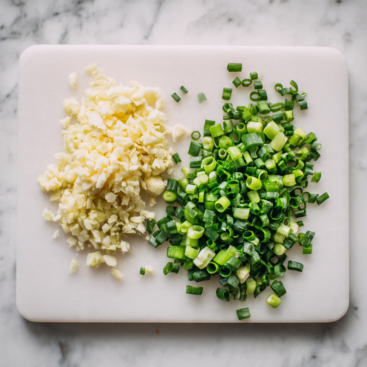 A white cutting board on a white marbled surface holds two piles: on the left, finely minced pale yellow garlic with a soft texture; on the right, chopped green onions in various shades of light and dark green, sliced into small round pieces and arranged in a loose pile. The smooth white cutting board contrasts with the vibrant colors of the ingredients. photo taken with an iphone --ar 4:5 --v 7