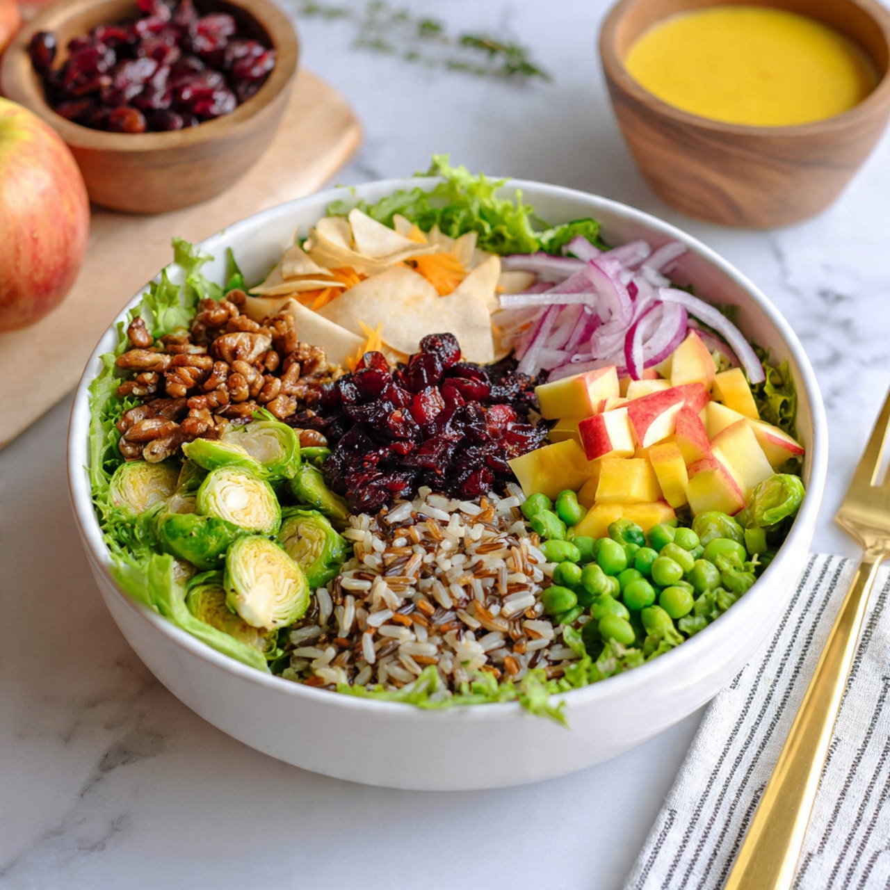 A white bowl sits on a white marbled surface, filled with seven layers of food arranged in sections. The bottom layer is a bed of green leafy lettuce. On the top left section, there are small, brown, crunchy nuts. Next to the nuts, thin slices of light beige root vegetable chips are layered on top of darker orange ones. In the center, there are thin, green slices of Brussels sprouts mixed with bits of dark red dried cranberries. To the right, a small mound of light and dark mixed brown rice grains creates a textured pile. Below the rice is a section of bright green peas. Near the bottom right, small yellow and red pieces of diced apple sit beside thin, curly slices of purple onions. On top of the peas and apples, shiny, dark red dried cranberries add a pop of deep color. In the background, there is a bowl of yellow dressing, a wooden bowl of dried cranberries, an apple, and part of another salad bowl. A gold fork and striped napkin rest on the right side of the frame. Photo taken with an iphone --ar 4:5 --v 7