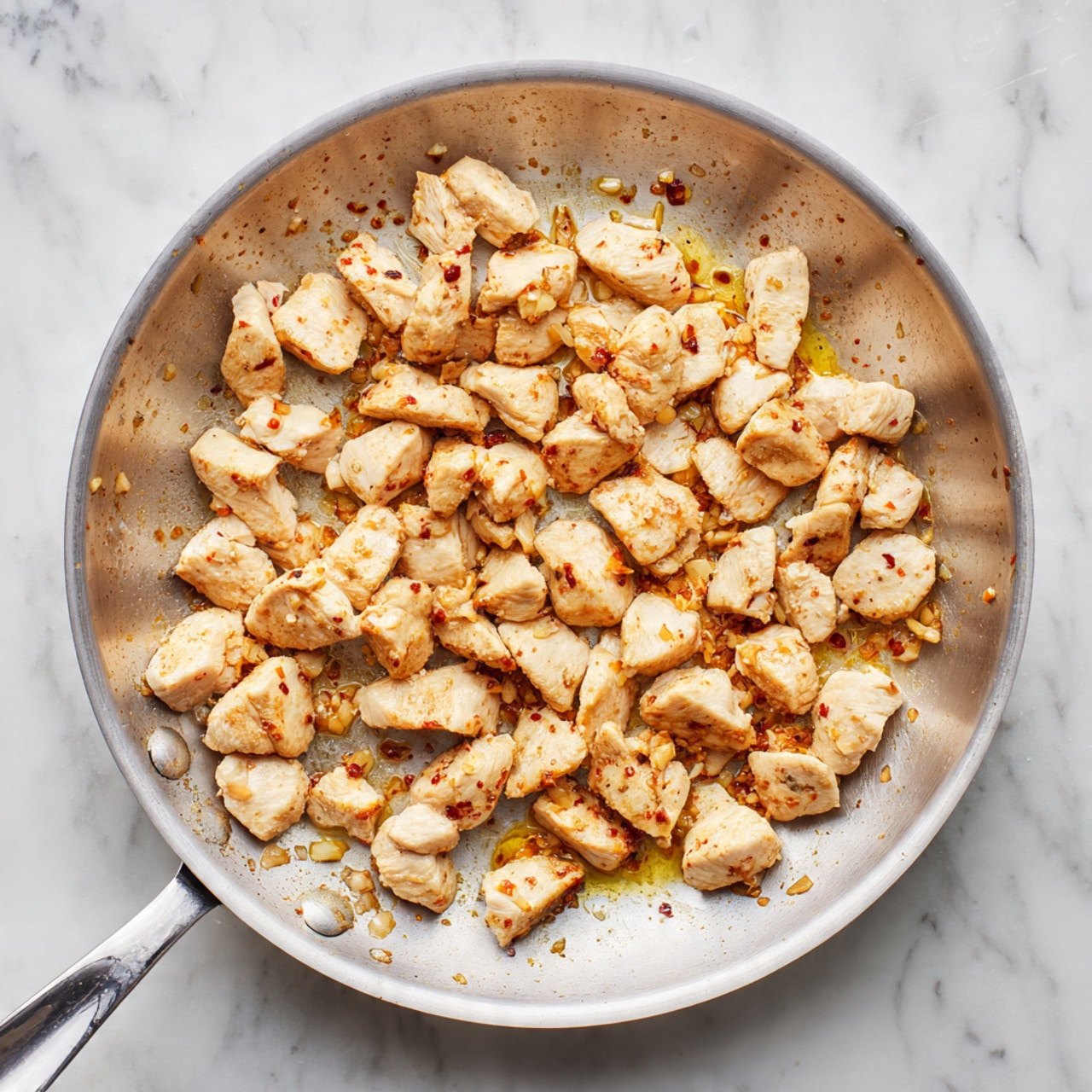 A silver frying pan on a white marbled surface holds many small pieces of cooked chicken. The chicken pieces are light beige with slight browning on some sides, scattered evenly across the pan. There are small spots of light golden oil and a few bits of cooked garlic around the chicken, giving a slightly shiny look. The pan has a smooth metal handle pointing to the lower left corner. photo taken with an iphone --ar 4:5 --v 7