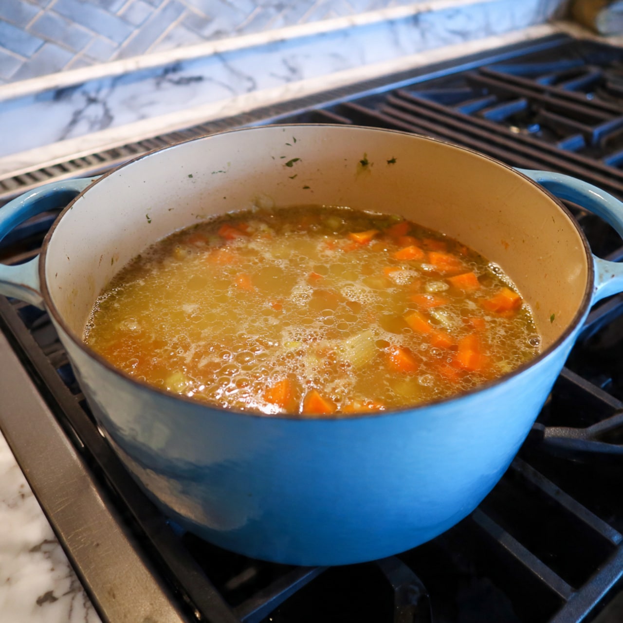 A large light blue pot sits on a stove with bubbling soup inside. The soup has several visible layers: a top layer of light foam bubbles, below that orange chunks of vegetables, likely sweet potatoes, floating in a clear brownish broth. The pot's inside is cream-colored, contrasting with the blue outside. The stove is dark with metal burner grates, and next to it is a white marbled countertop. photo taken with an iphone --ar 4:5 --v 7