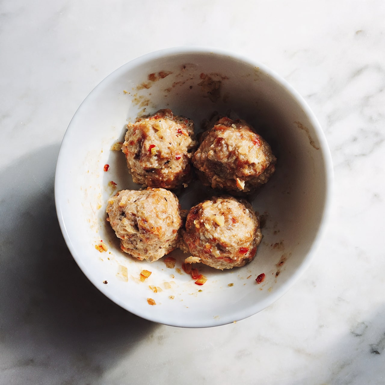 The image shows a white bowl filled with four round meatballs, placed evenly inside the bowl. The meatballs have a coarse texture with visible bits of white and red throughout, indicating mixed ingredients. The inside of the bowl has small bits and smudges of the meat mixture. The bowl sits on a white marbled surface. photo taken with an iphone --ar 4:5 --v 7
