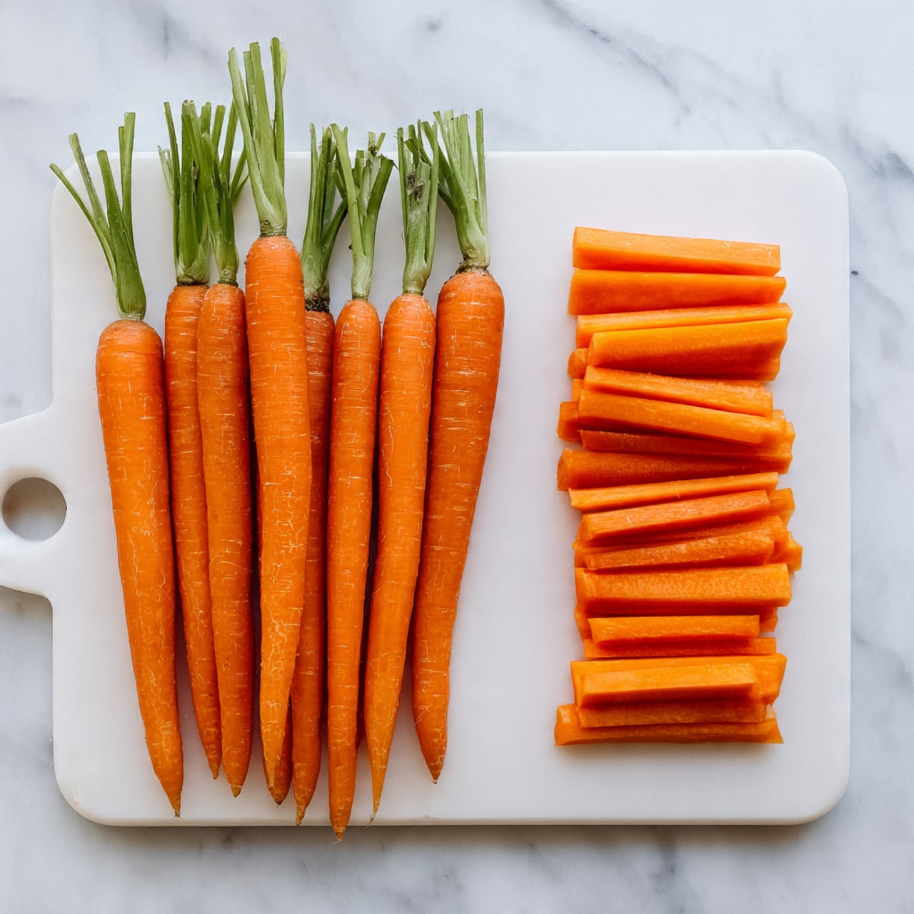 The image shows a white cutting board on a white marbled surface with fresh bright orange carrots. On the left side, there are seven whole carrots with green tops removed, arranged in a slightly fanned out way. On the right side, one carrot is cut into many short thick pieces, all neatly lined up horizontally. The carrots have a smooth texture and vibrant color. photo taken with an iphone --ar 4:5 --v 7