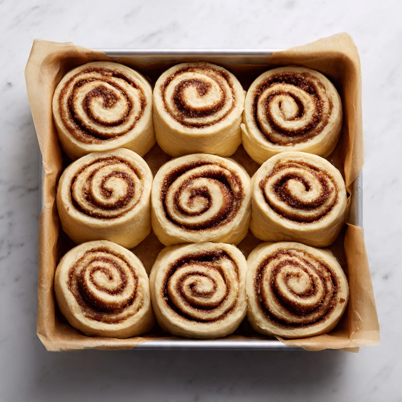 A metal baking pan lined with light brown parchment paper holds twelve unbaked cinnamon rolls arranged in three rows of four. Each cinnamon roll has a spiral shape with layers of light dough and darker cinnamon filling visible, showing a rough texture. The rolls are evenly spaced with some dough edges slightly thicker and pale cream in color. The background is a white marbled surface. photo taken with an iphone --ar 4:5 --v 7