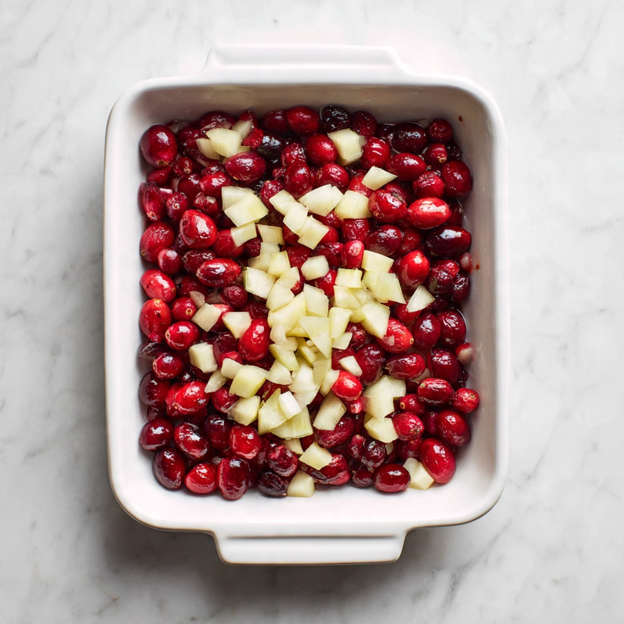 The image shows a white rectangular baking dish filled with two layers of food. The bottom layer is made of whole and halved dark red cranberries, creating a deep red base with a glossy texture. On top, there is a scattered layer of small, pale yellow diced pieces that look like fruit or vegetables, placed evenly across the cranberries. The dish sits on a white marbled surface, and natural light highlights the freshness and color contrast of the ingredients. Photo taken with an iphone --ar 4:5 --v 7