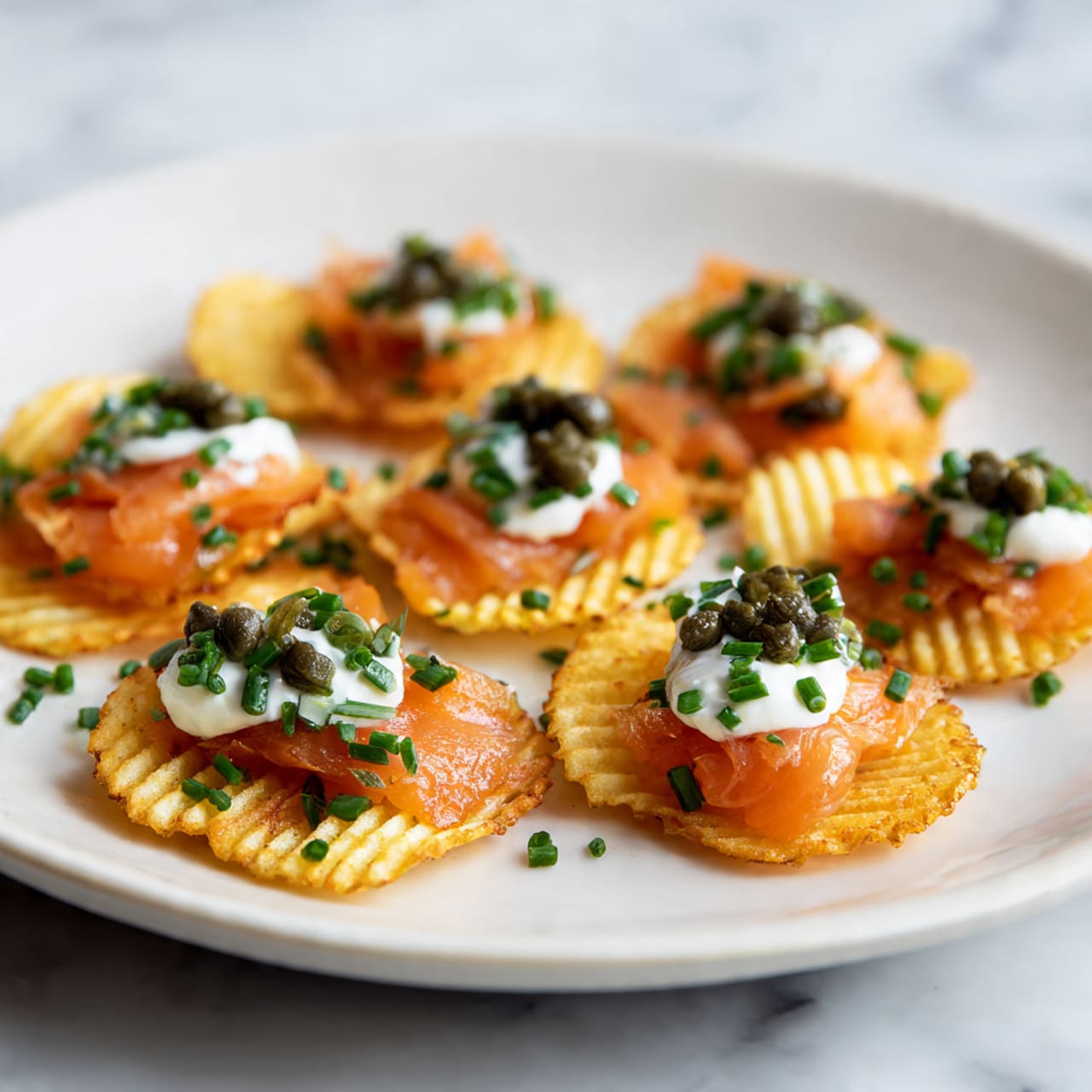 This image shows a white plate filled with small round ridged potato chips as the base layer. On each chip, there is a thin layer of orange smoked salmon folded softly in the middle layer. On top of the salmon, there is a small dollop of white cream. The cream is garnished with small green chopped chives and dark green capers, placed evenly across the plate. The plate is set on a white marbled surface. photo taken with an iphone --ar 4:5 --v 7